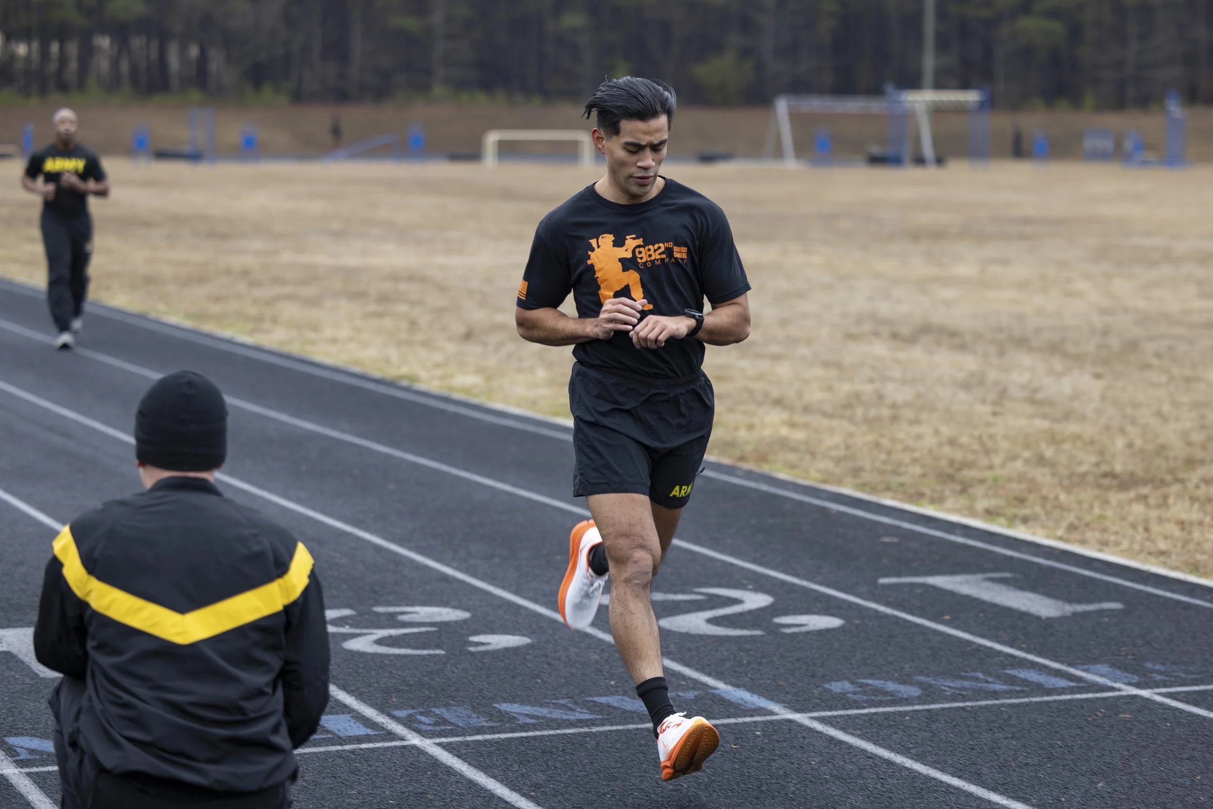 A young man running on a track during daytime, with a serious expression, wearing a black athletic outfit and orange running shoes. There is a person in a black jacket and hat sitting near the track, and another person walking in the background.