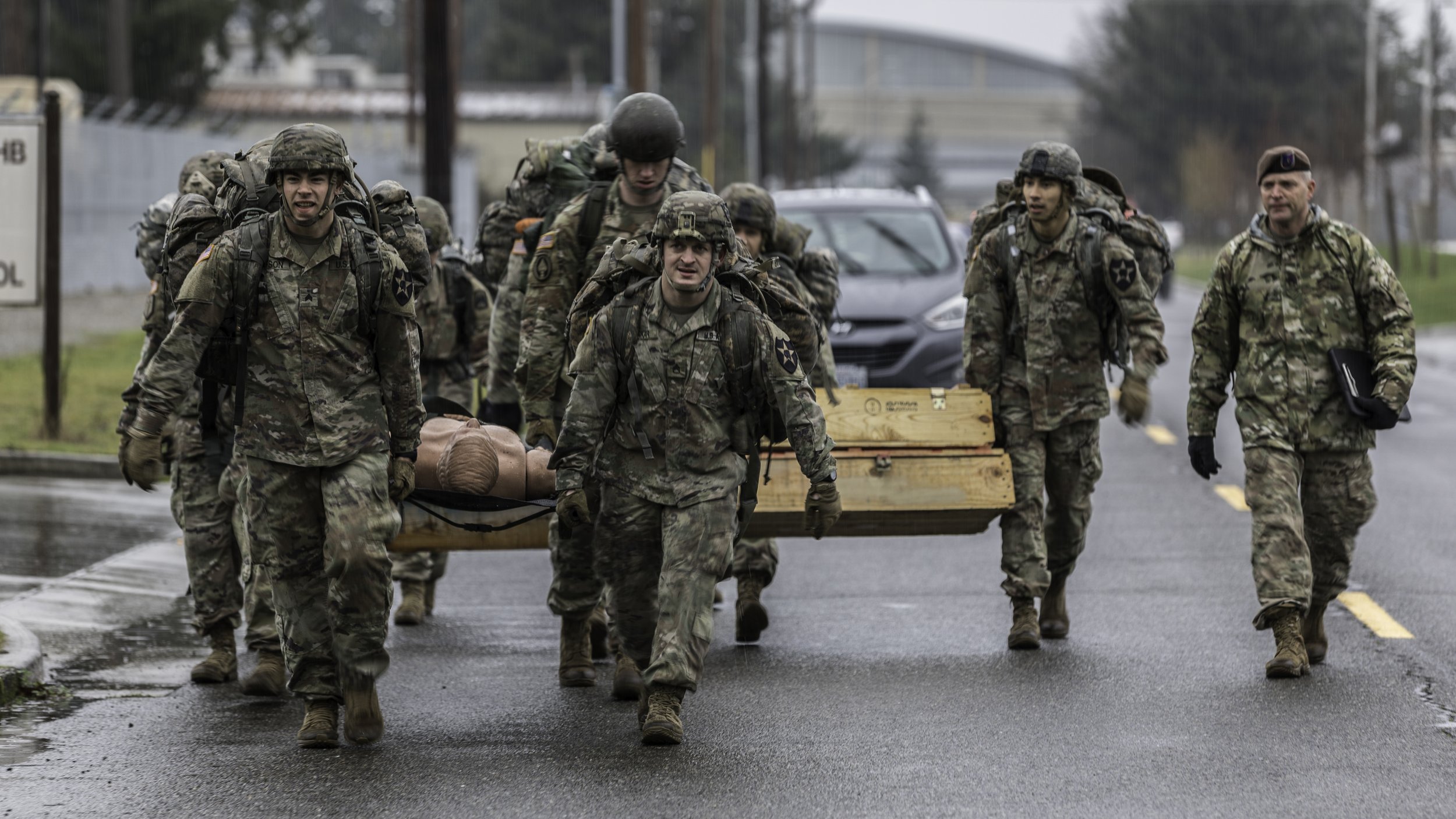 Group of soldiers in camouflage uniforms and helmets carrying a wounded person on a stretcher on a wet street.
