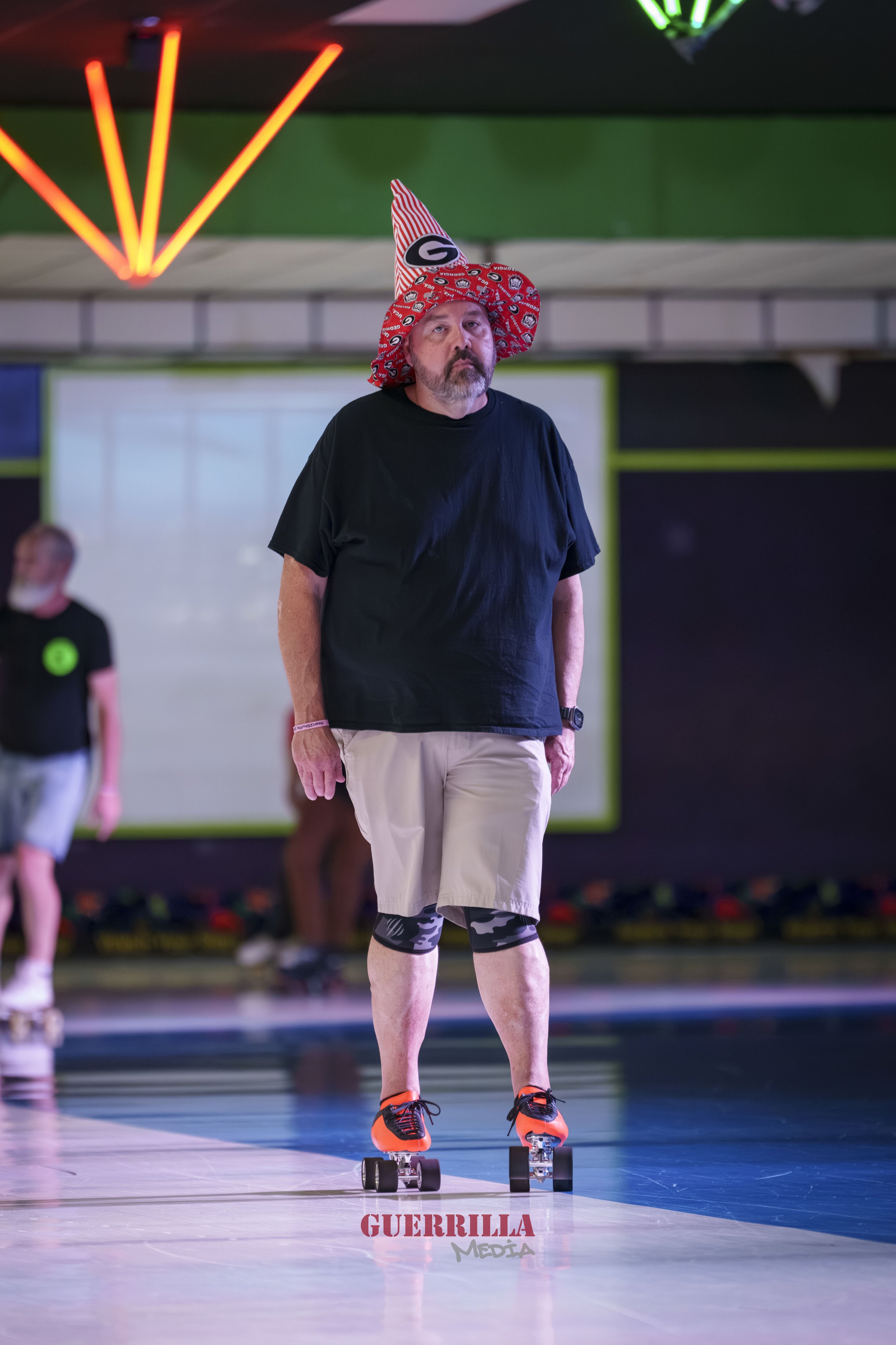 A man wearing a black T-shirt, beige shorts, black patterned knee sleeves, and orange skate shoes, riding a skateboard on an indoor skating rink. He has a red and white party hat with a green 'G' logo and a red bandana pattern. The background feature