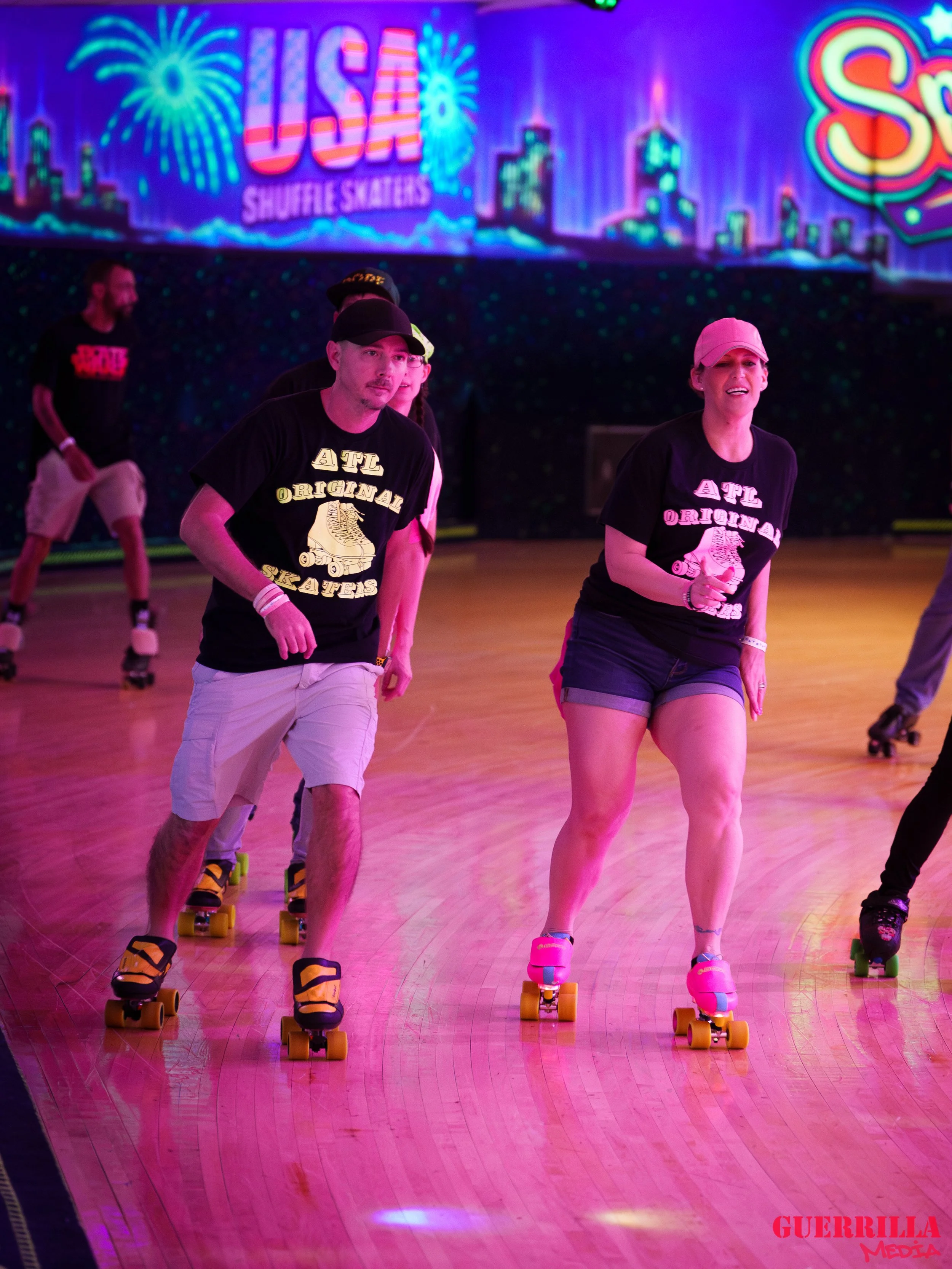 People roller skating under colorful neon lights at a roller skating rink with a backdrop that says 'USA Shuffle Skaters' and features fireworks and city skyline illustrations.