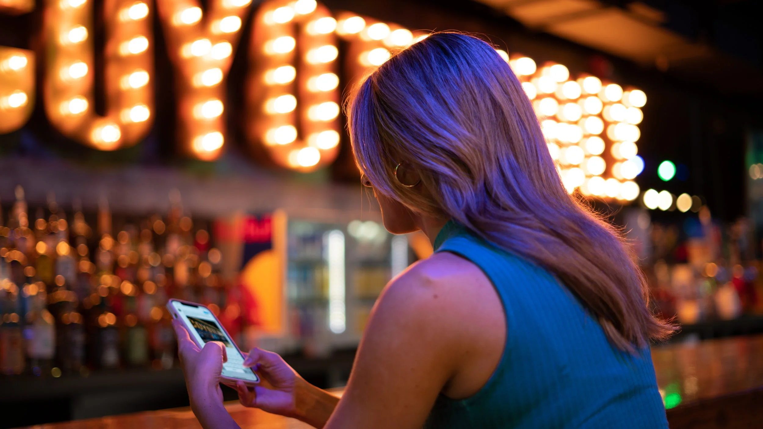 A woman with shoulder-length hair wearing a sleeveless blue top is using her smartphone at a bar or nightclub with colorful lights and blurred bottles in the background.