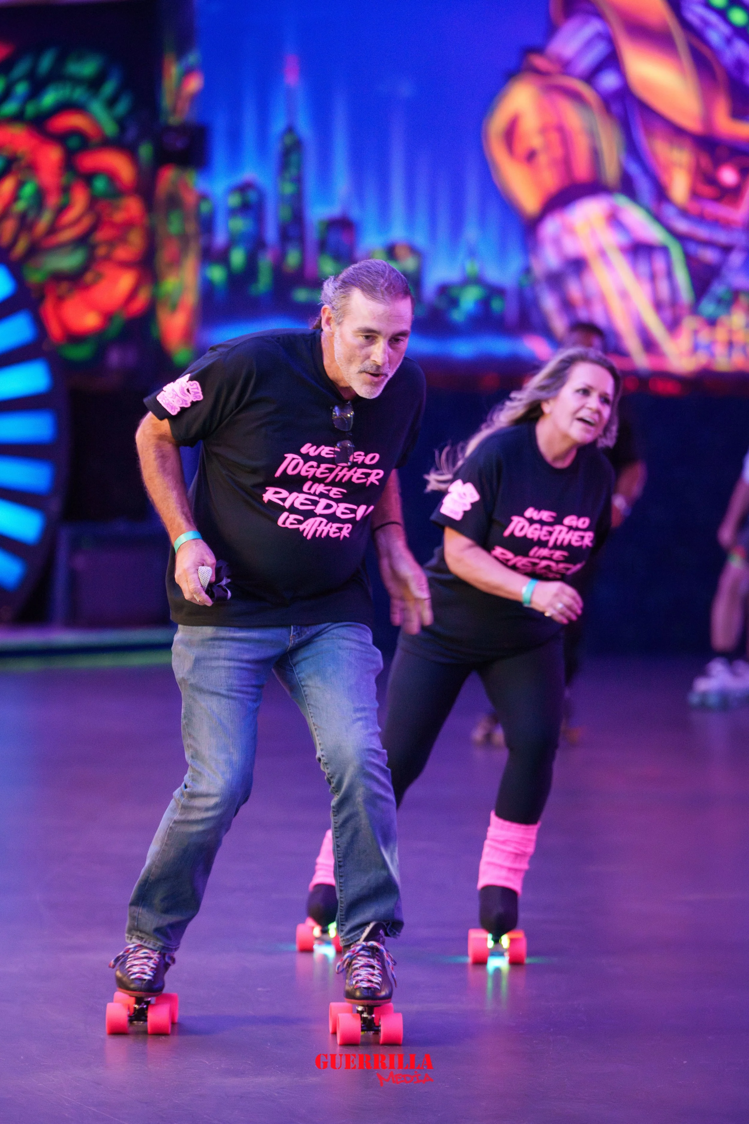 Two people roller skating, wearing matching black t-shirts with pink text, in a neon-lit indoor roller rink with colorful graffiti-style wall art.