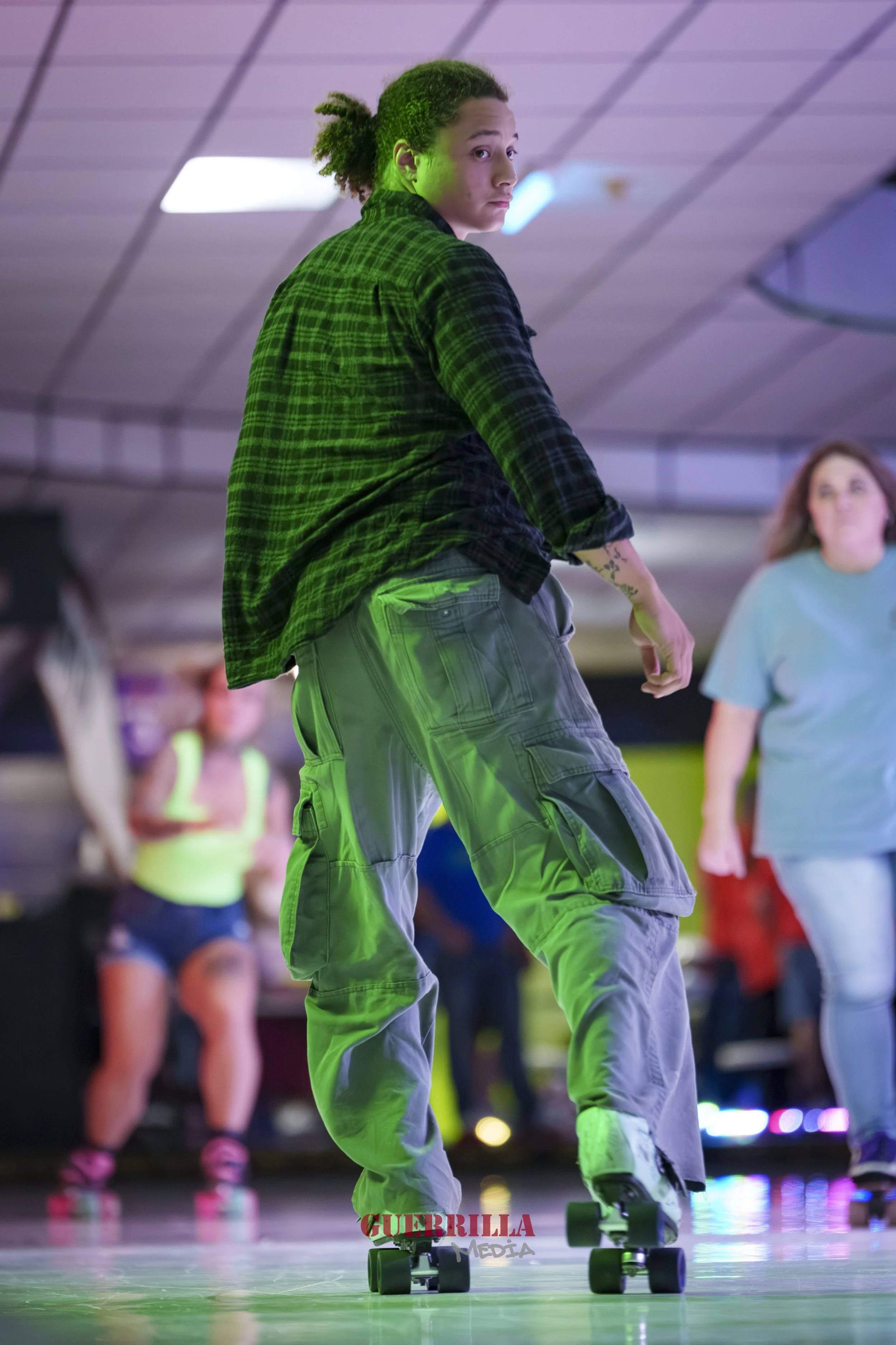 A person with curly hair skating indoors with other people in the background, illuminated by colorful lights.