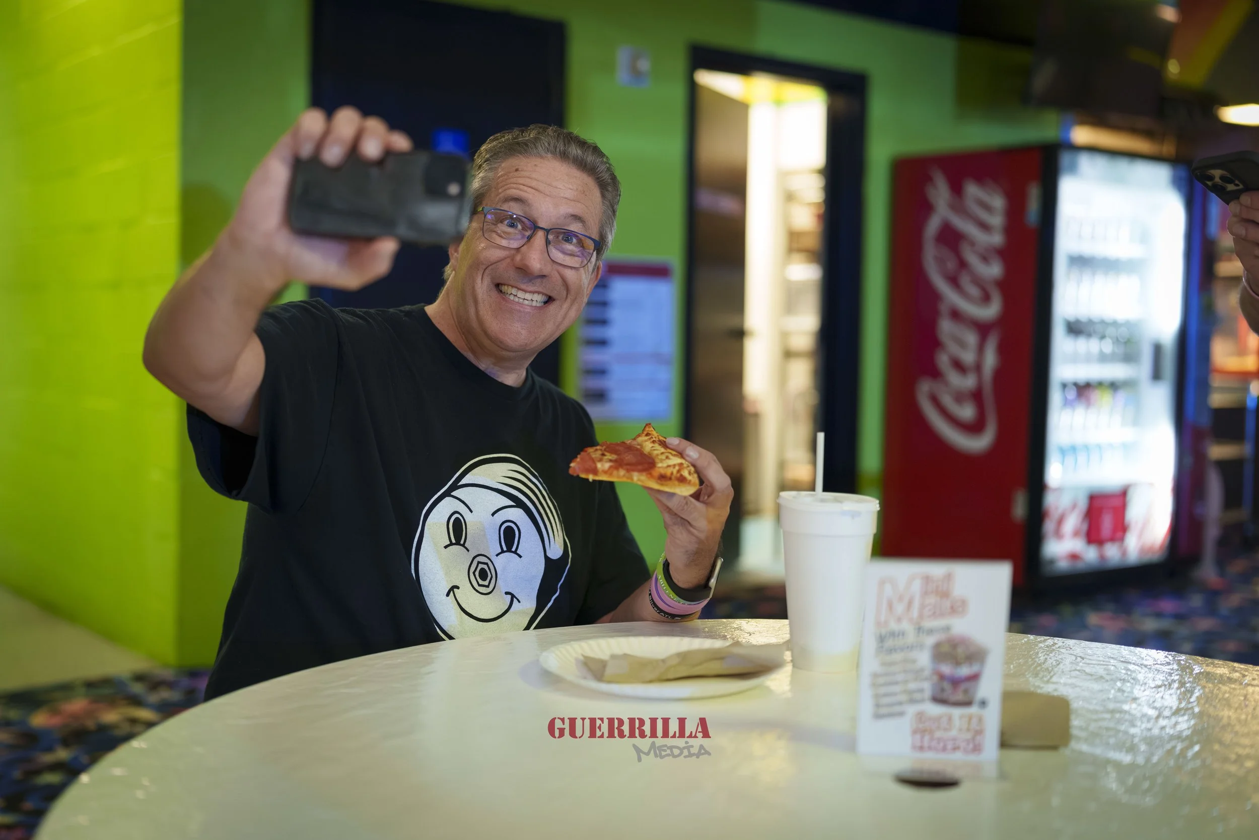 A smiling man with glasses, wearing a black t-shirt with a smiley face, is taking a selfie while holding a slice of pizza in a casual setting. There is a soda cup and a small menu on the table, with bright green walls and vending machines in the back