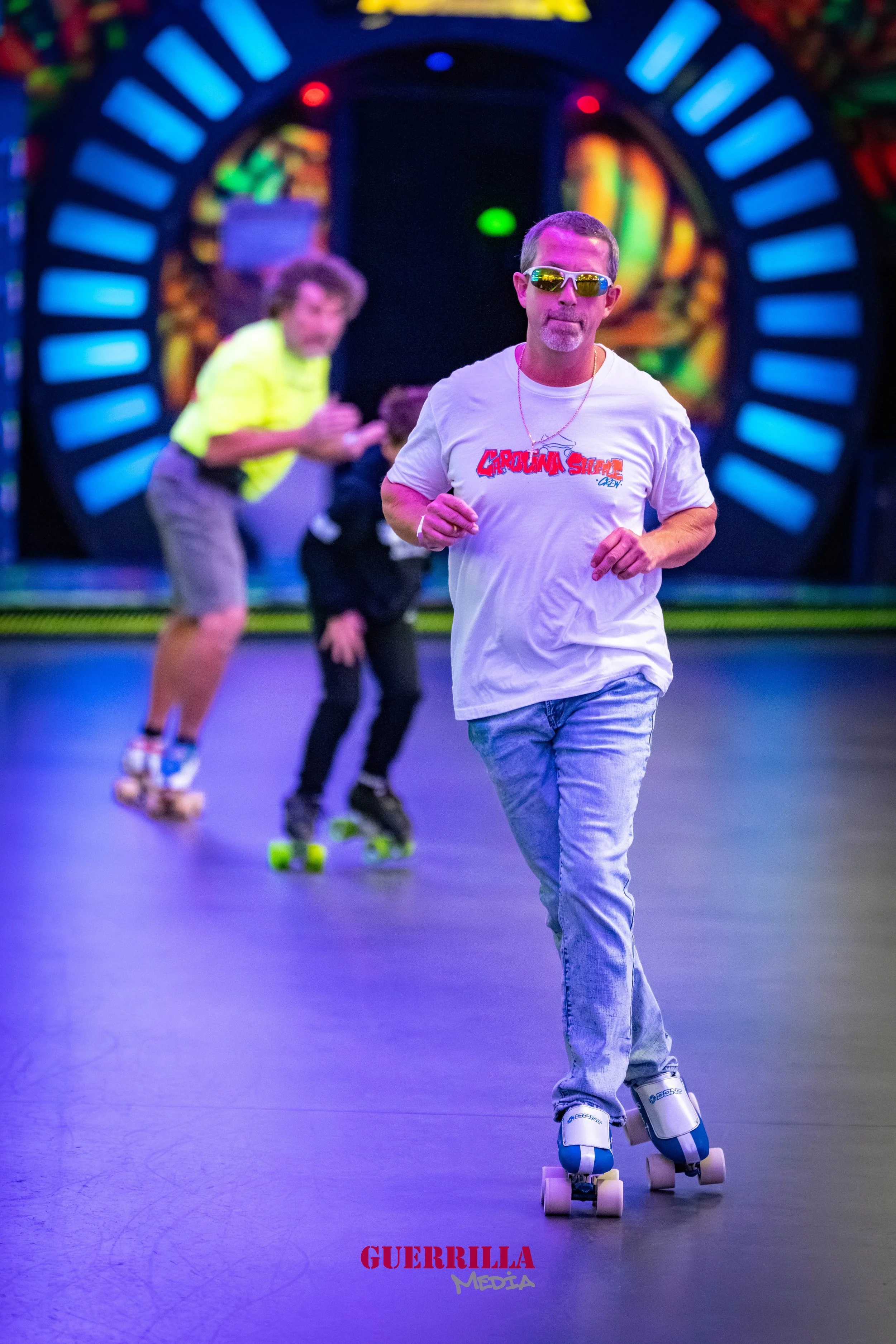 A man wearing sunglasses, a white T-shirt with red and blue text, and light jeans roller-skating in an indoor skating rink. Two children are skating behind him in the background, with one wearing a neon yellow shirt. The background features colorful,