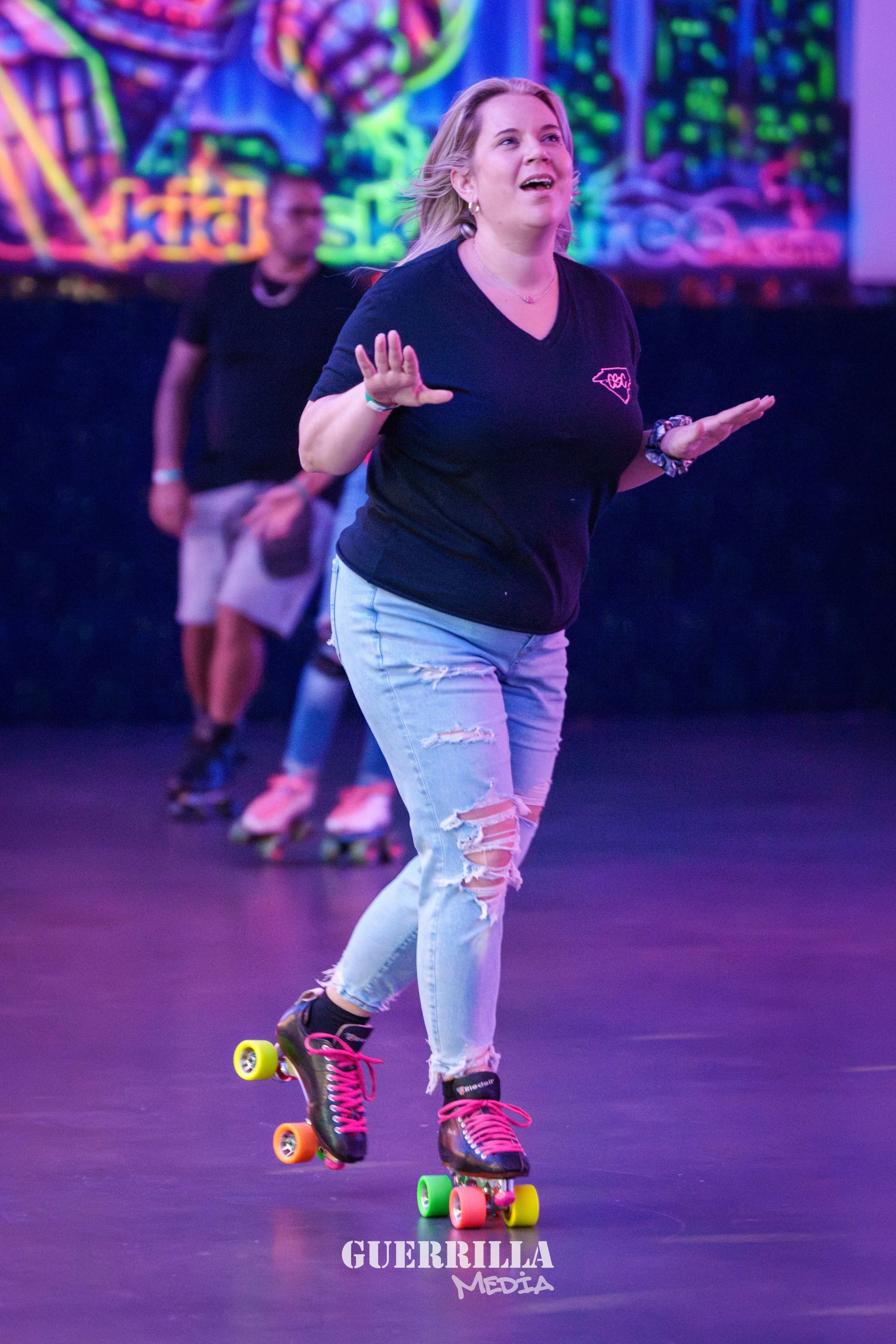 Woman roller skating indoors, wearing a black t-shirt with a pink logo, ripped light blue jeans, and pink laces. She is holding her hands up in a balancing posture, with a background of illuminated neon signs and people also skating.