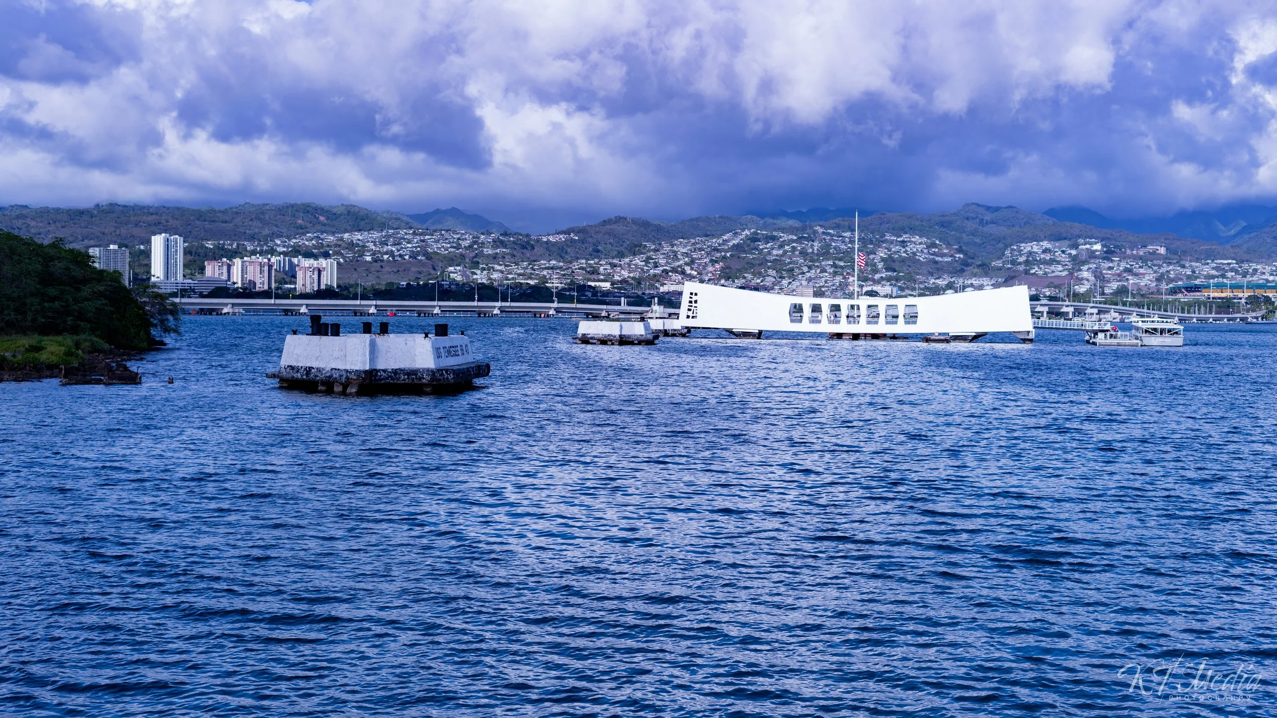 View of a harbor with a submerged white structure that looks like a ship's bow, an American flag flagpole, and a city with high-rise buildings and mountains in the background. Overcast sky with patches of blue.