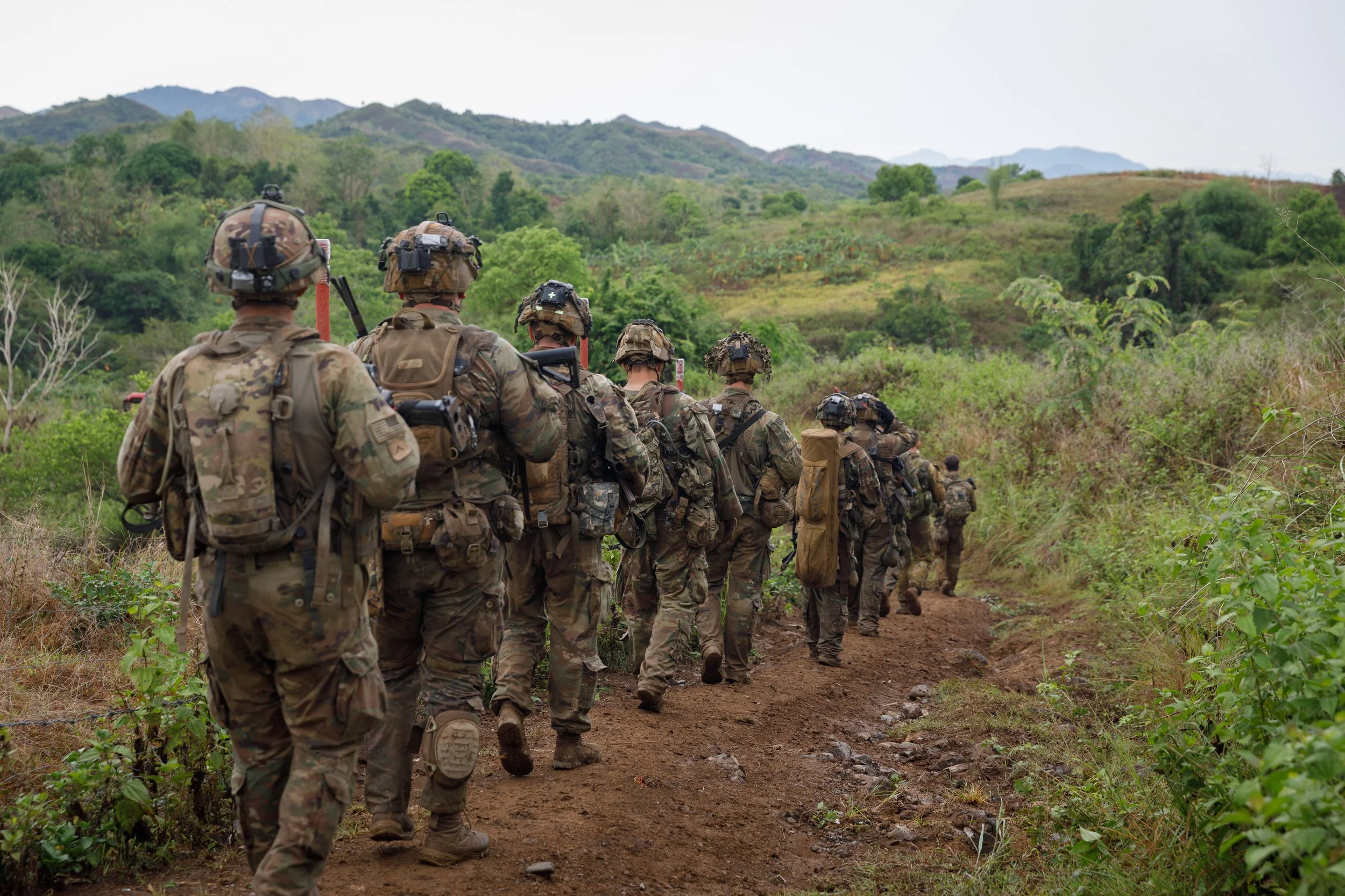 A group of soldiers in camouflage uniforms walking in a line on a dirt trail through a green, hilly landscape.