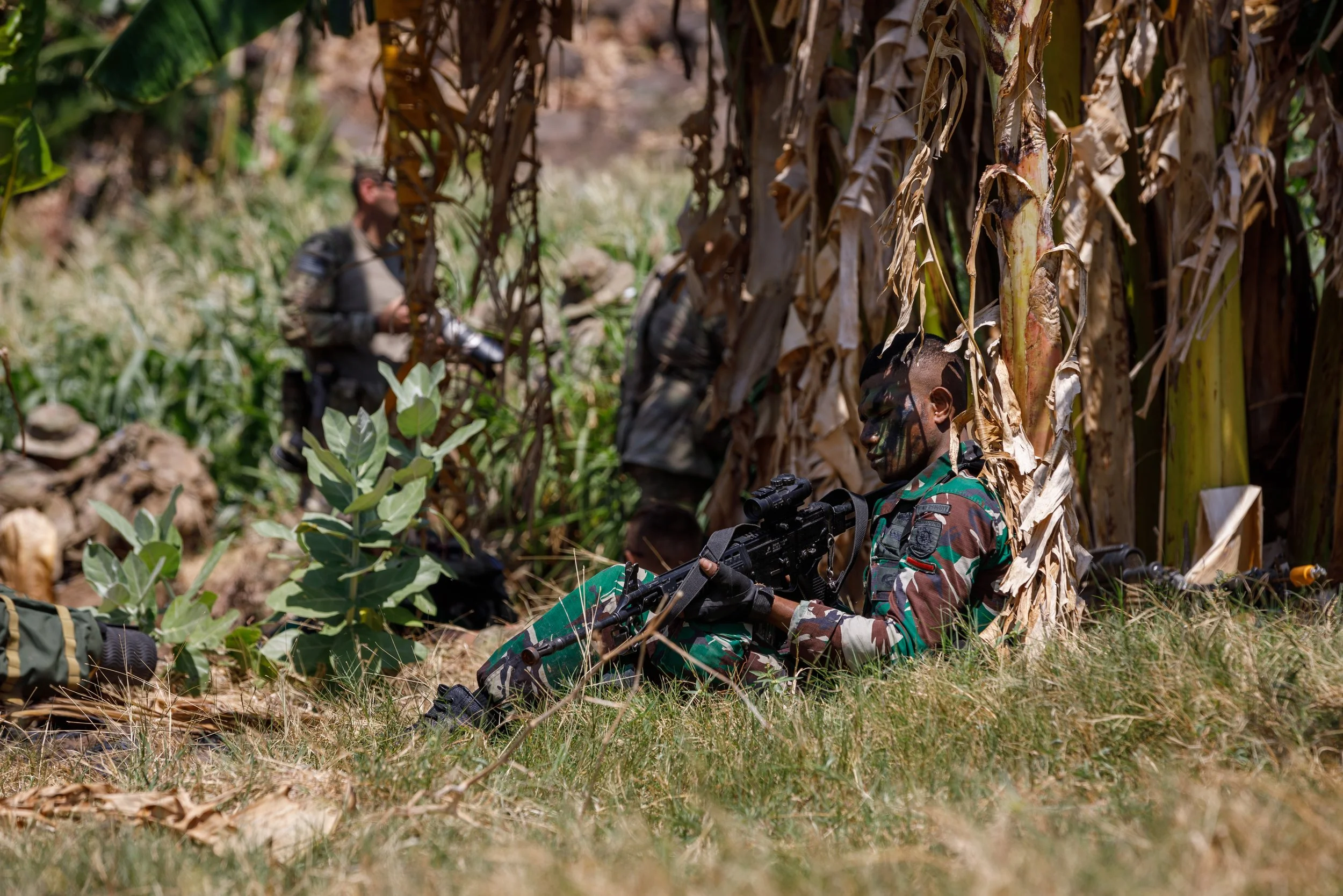 Soldiers in camouflage uniforms resting in a field of tall grass and banana trees, with one soldier sitting against a tree holding a sniper rifle.