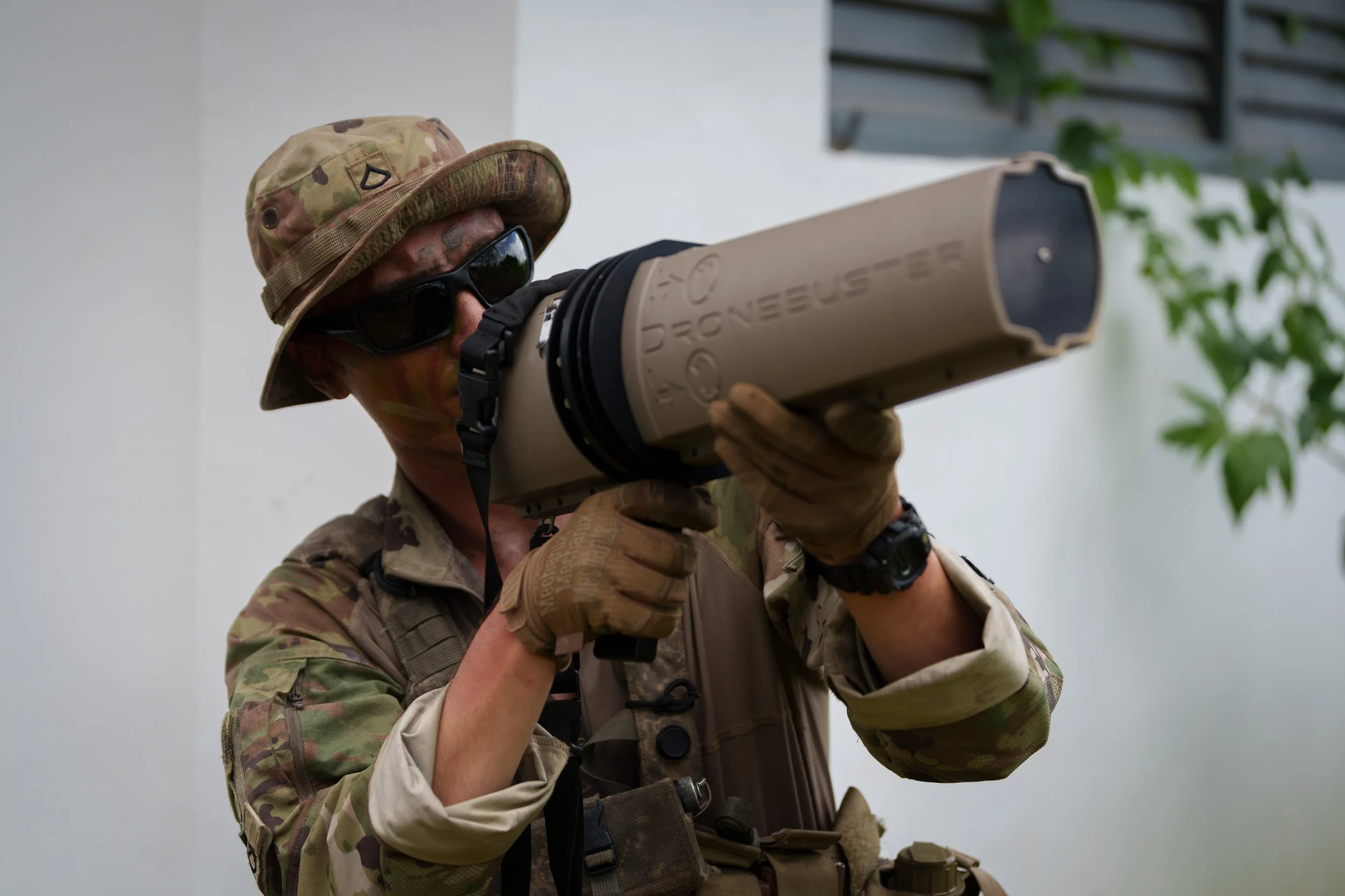 A soldier in camouflage uniform, wearing a boonie hat and sunglasses, holding a 'Dronesbuster' aiming it upward.