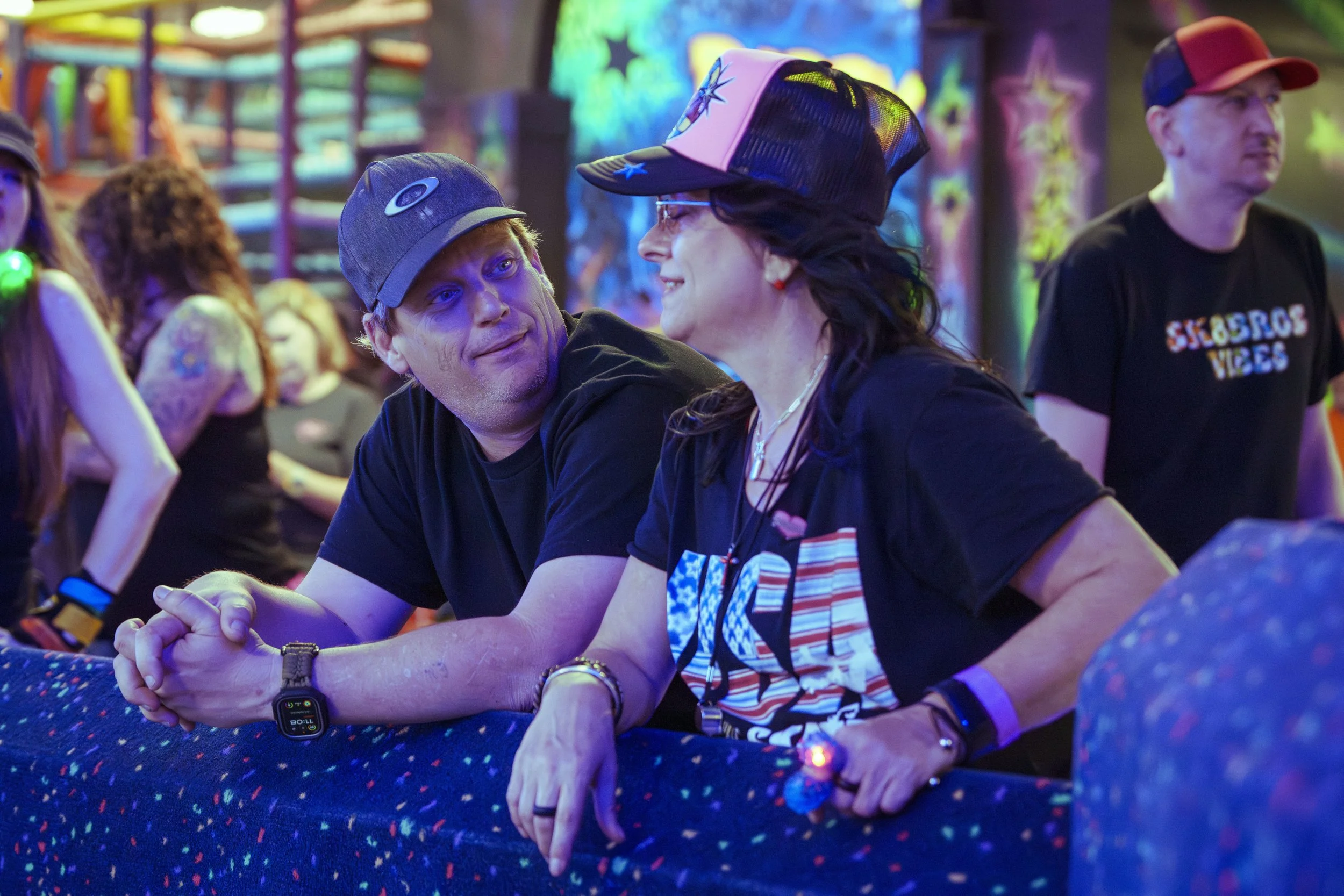 Two people, a man and a woman, talking at an indoor event or concert with vibrant, neon-colored lights. The man is wearing a black t-shirt and a cap, while the woman is wearing a black t-shirt and a baseball cap. They are leaning on a railing, engagi