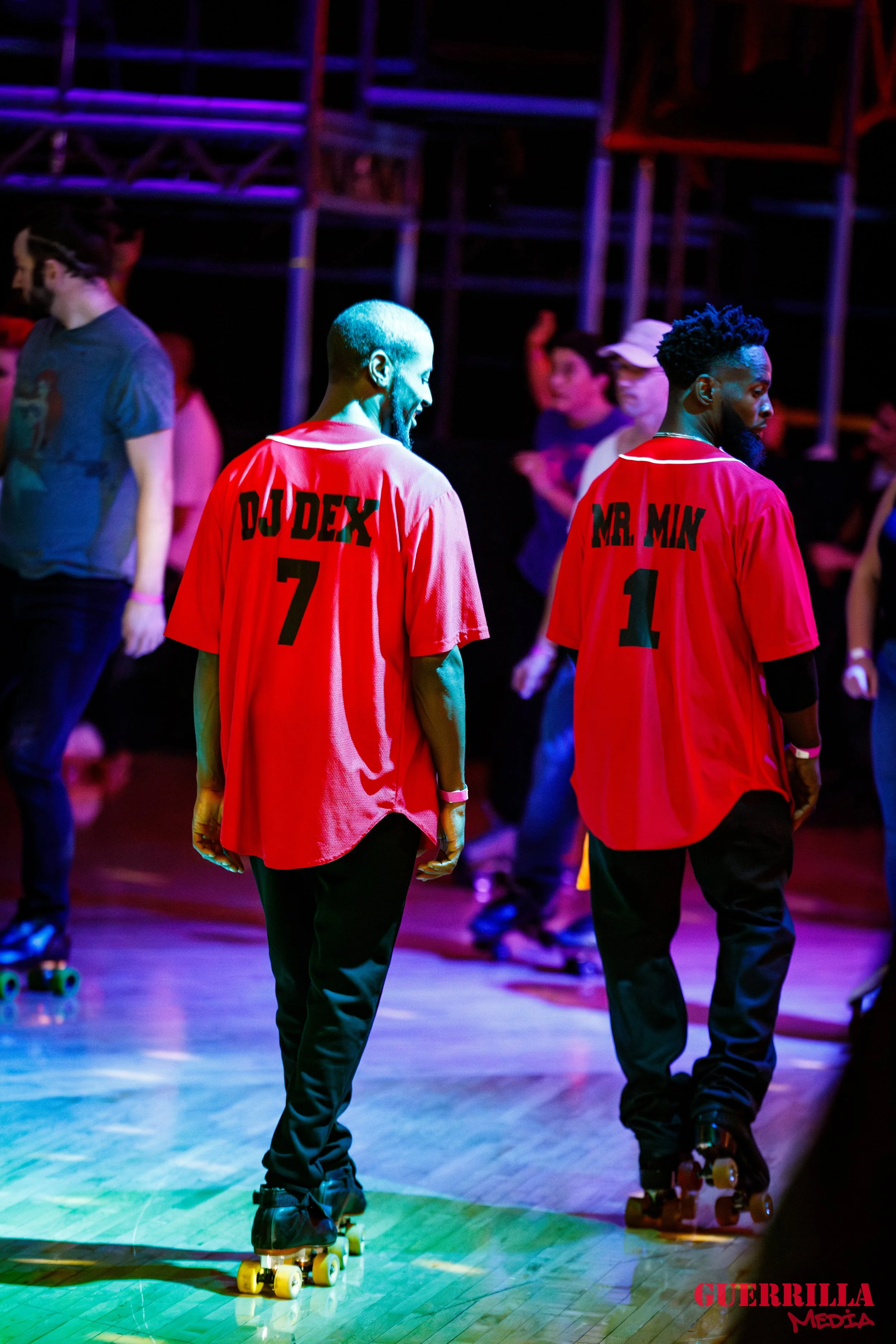 Two men roller skating at a dance event, wearing red shirts with black text on the back reading 'DJ DEX 7' and 'MR. MIN 1'. They are surrounded by other skaters and a dark background with colorful lighting.