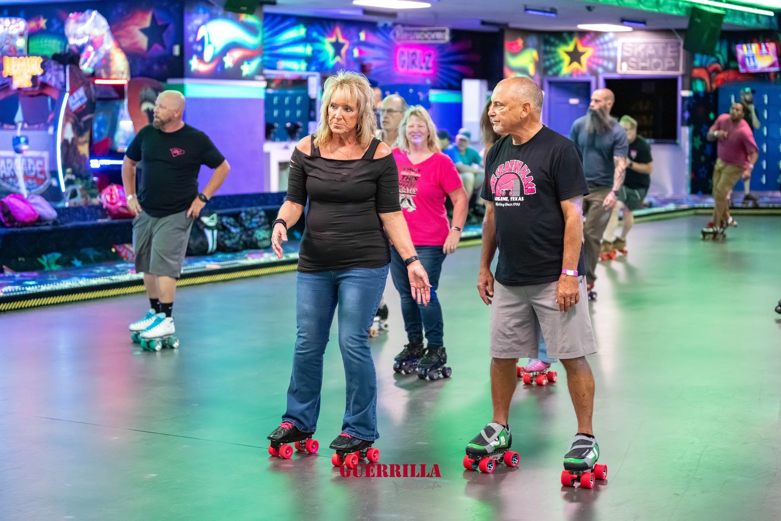 People roller skating indoors at a colorful roller rink with neon lights, including a woman and a man in the foreground engaged in conversation.