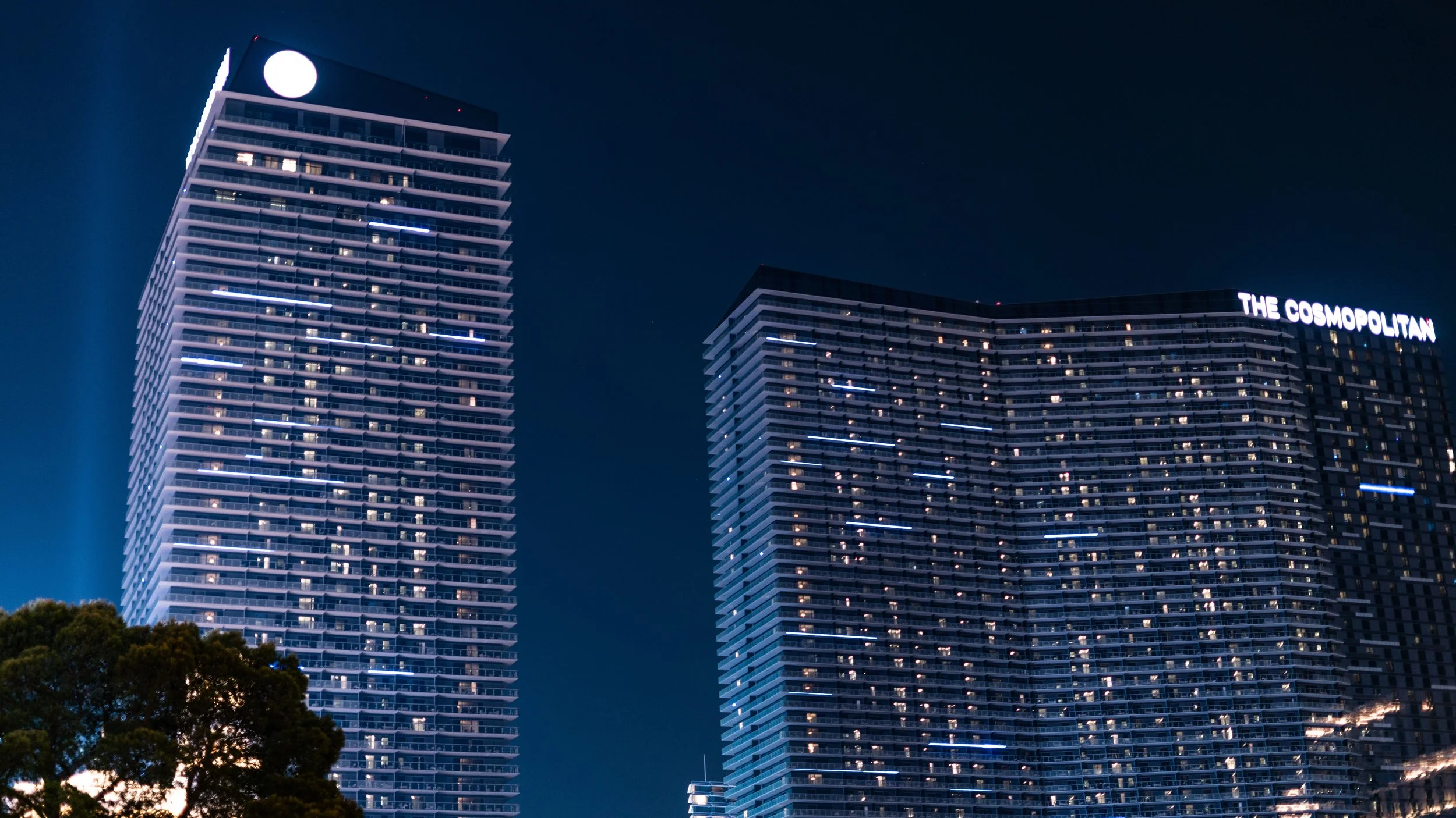 Night view of two tall skyscrapers with illuminated windows, one with a round light on top and the other with 'The Cosmopolitan' sign on it. Trees are visible in the foreground.
