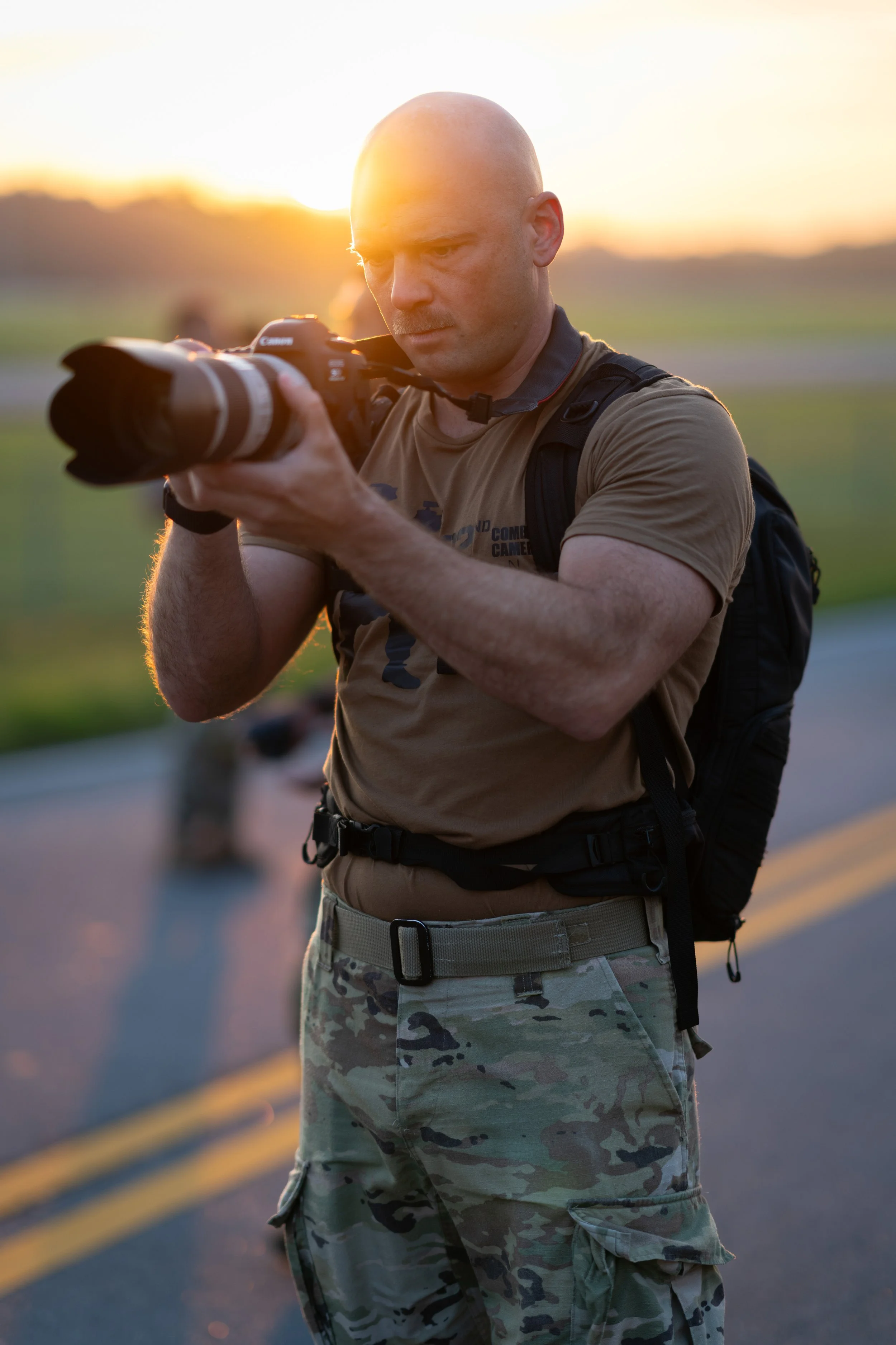 A man with a shaved head wearing a tan T-shirt and camouflage cargo pants is holding a professional camera with a large lens, aiming through the viewfinder against the backdrop of a sunset with a rural landscape.