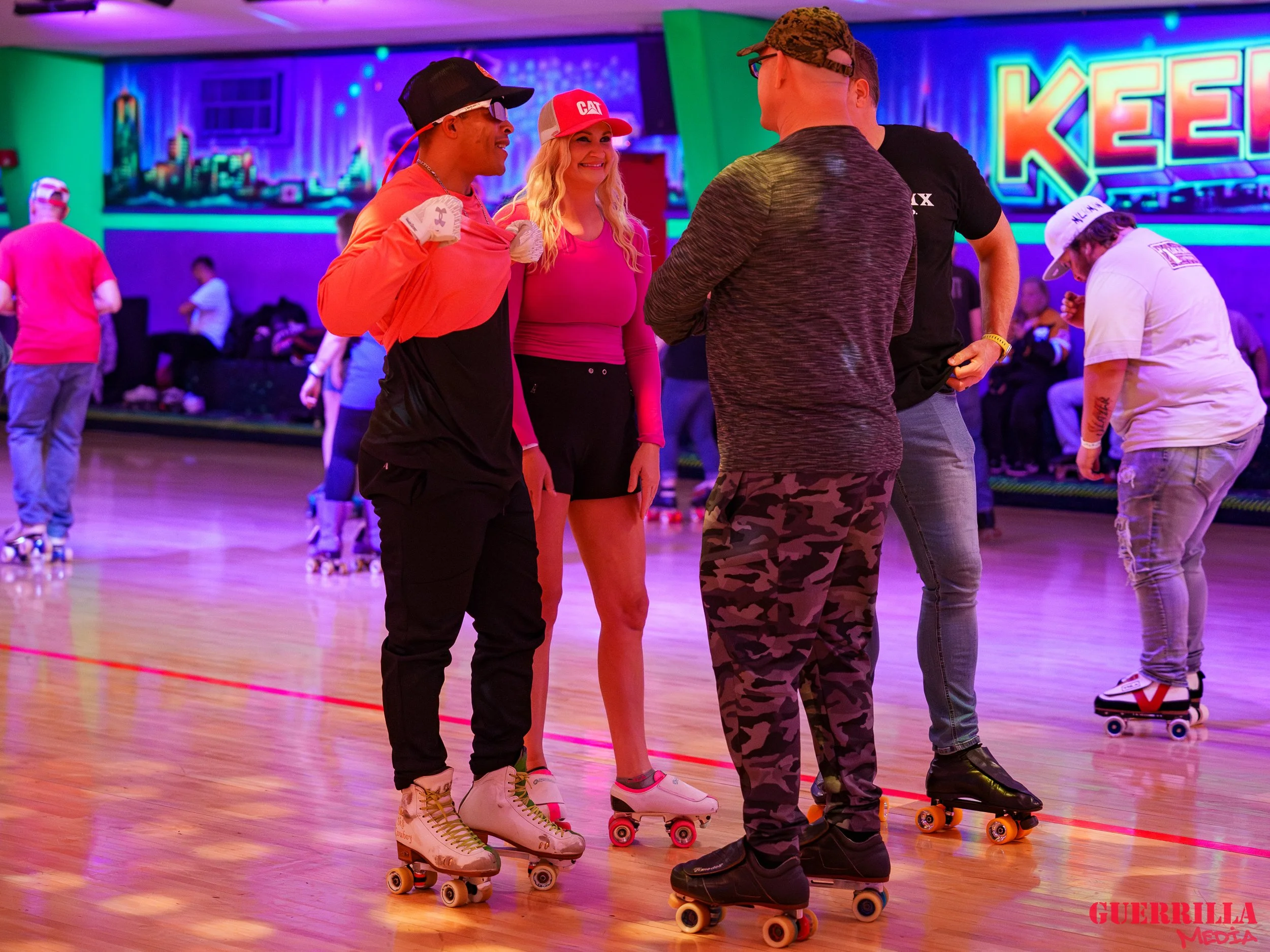 Group of people roller skating in an indoor roller rink with colorful neon lights, including three men and one woman in the foreground, with others skating and sitting along the wall in the background.