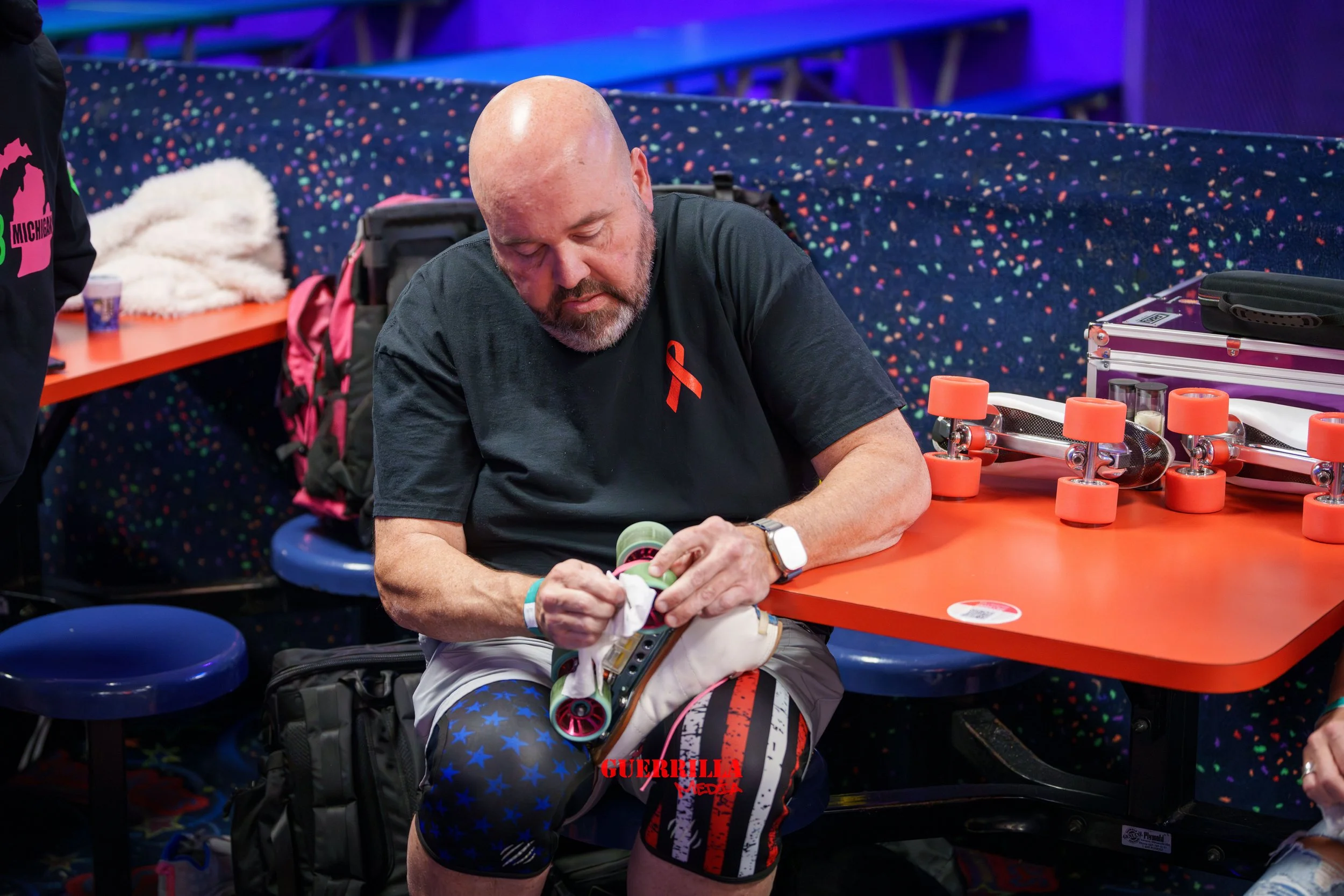 A man with a shaved head and gray beard sitting at a roller skating rink, applying tape to his roller skate. He wears a black T-shirt with an orange ribbon, and patterned shorts with American flag elements. There are orange skate wheels and a purple 