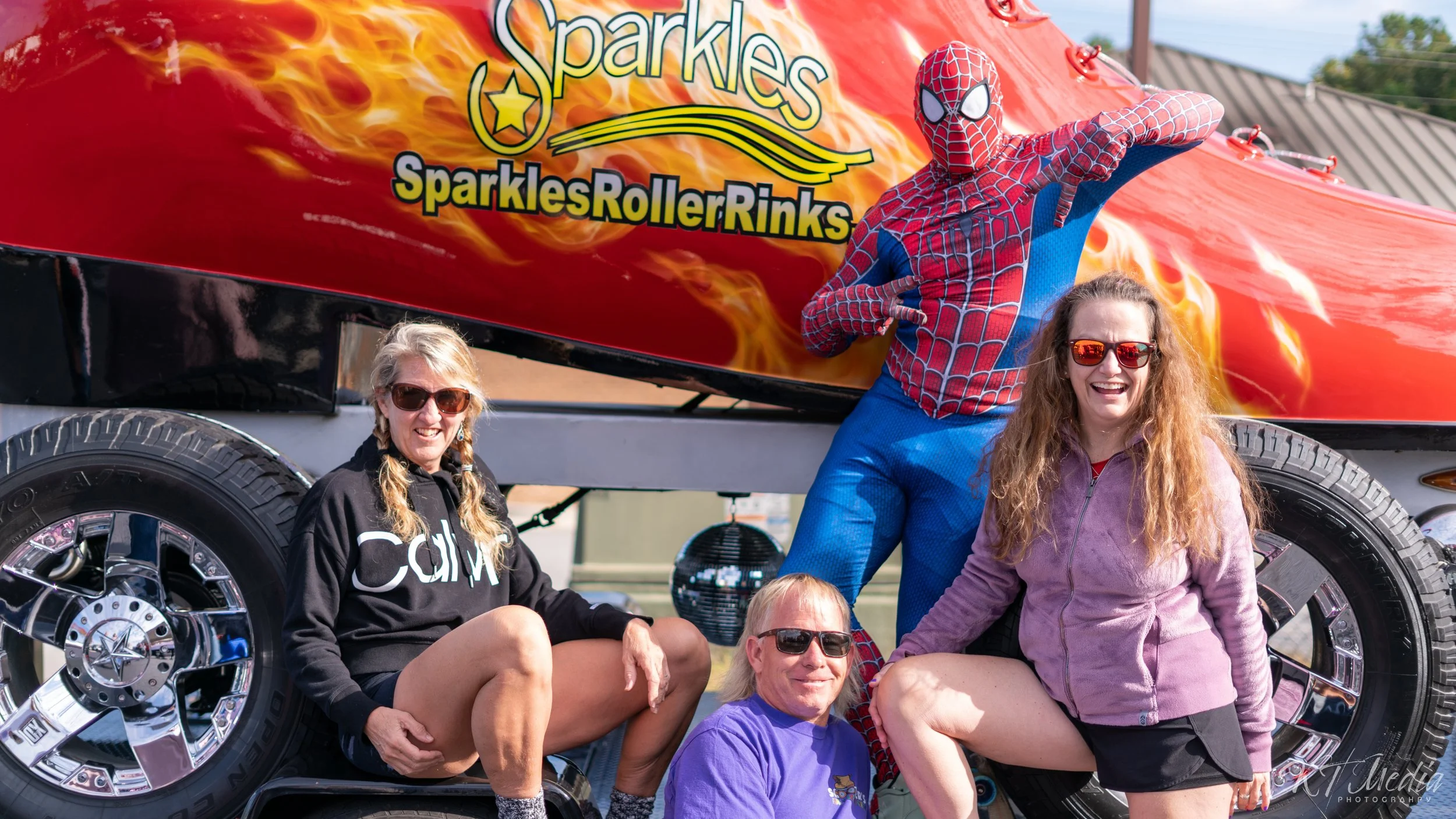 Group of four women posing in front of a red vehicle with a Spider-Man mascot, at an outdoor event.