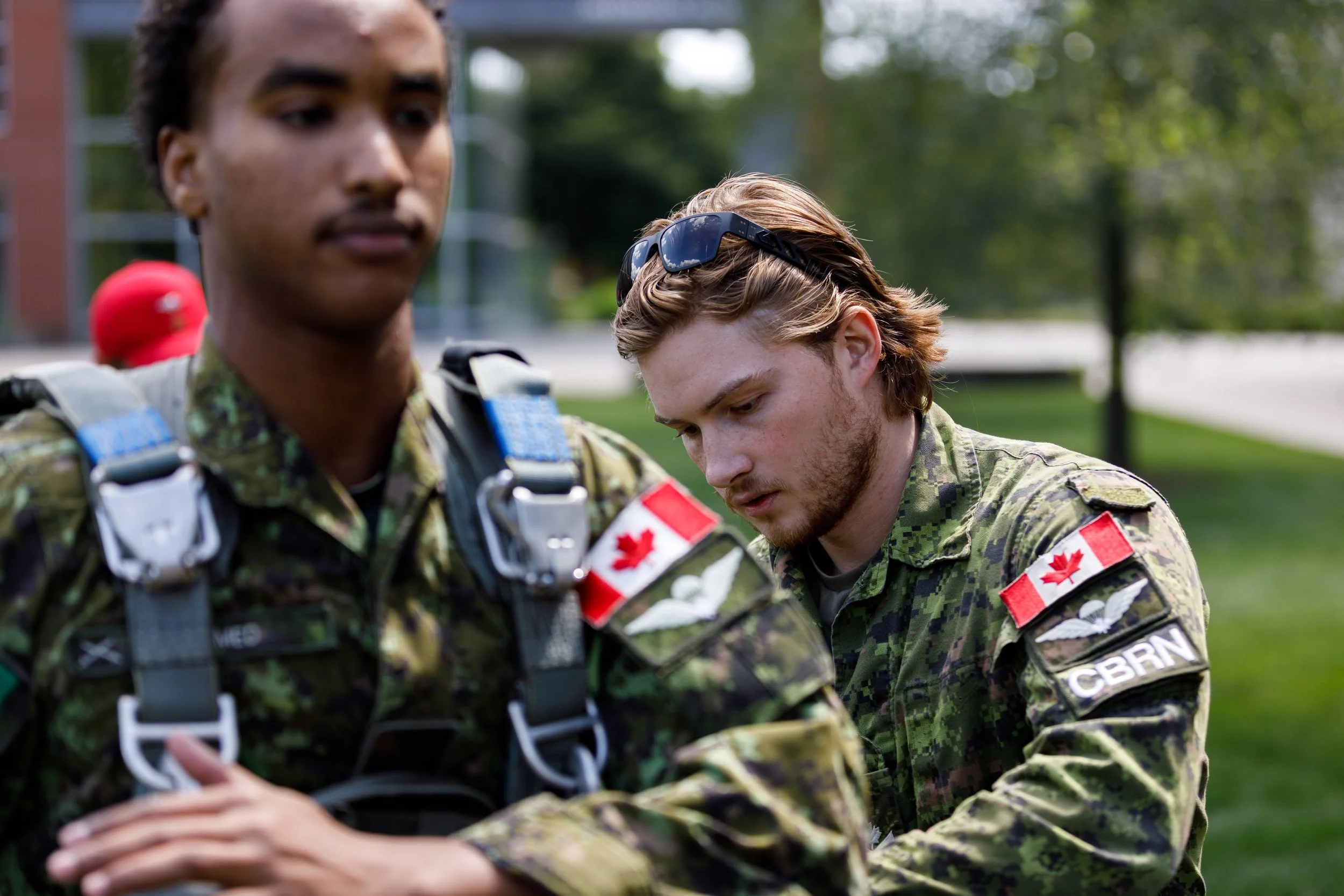 Two soldiers in camouflage uniforms with Canadian flags on their shoulders, one standing with arms crossed and the other adjusting gear, outdoors with trees in the background.