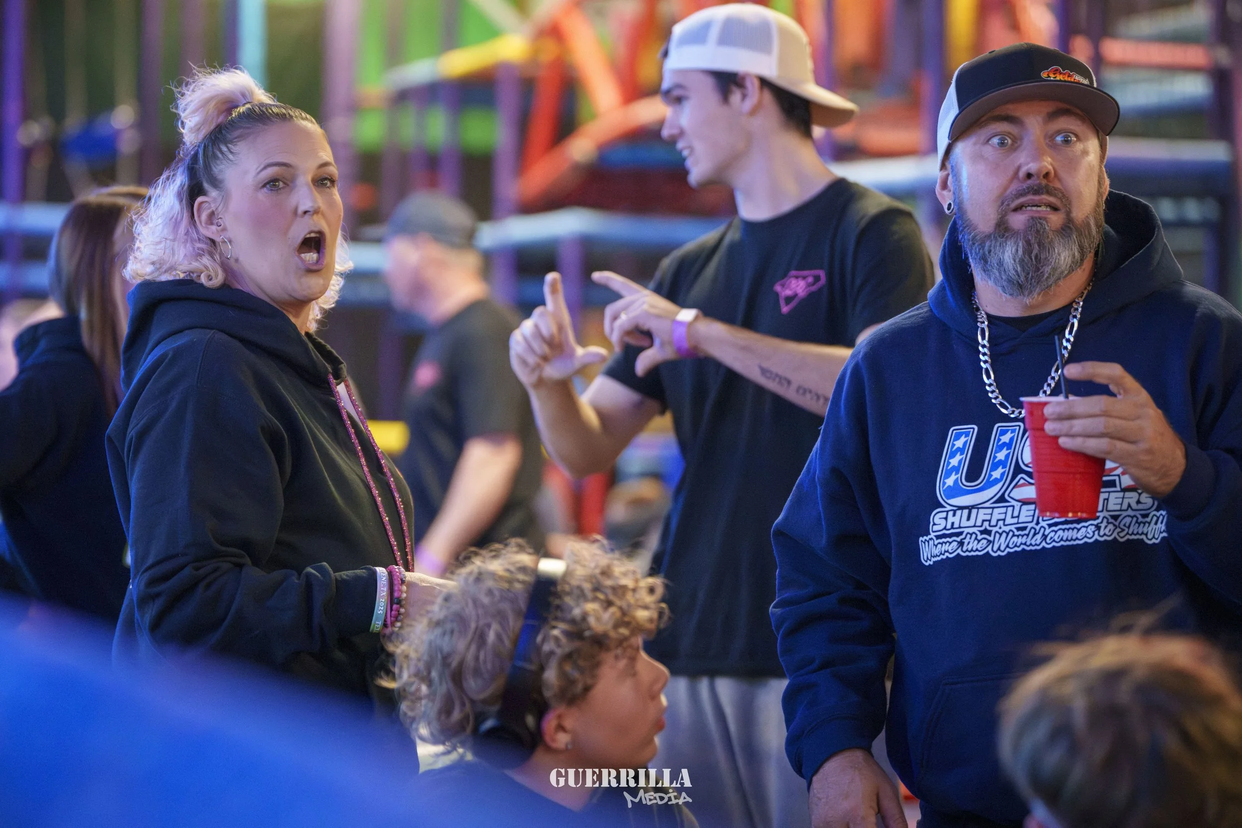 A group of people at a colorful amusement park or carnival. A woman with pink hair styled in a bun is talking with her mouth open, wearing a black hoodie. A man with a beard, wearing a black hoodie, a silver chain, and a black cap, holds a red cup. A