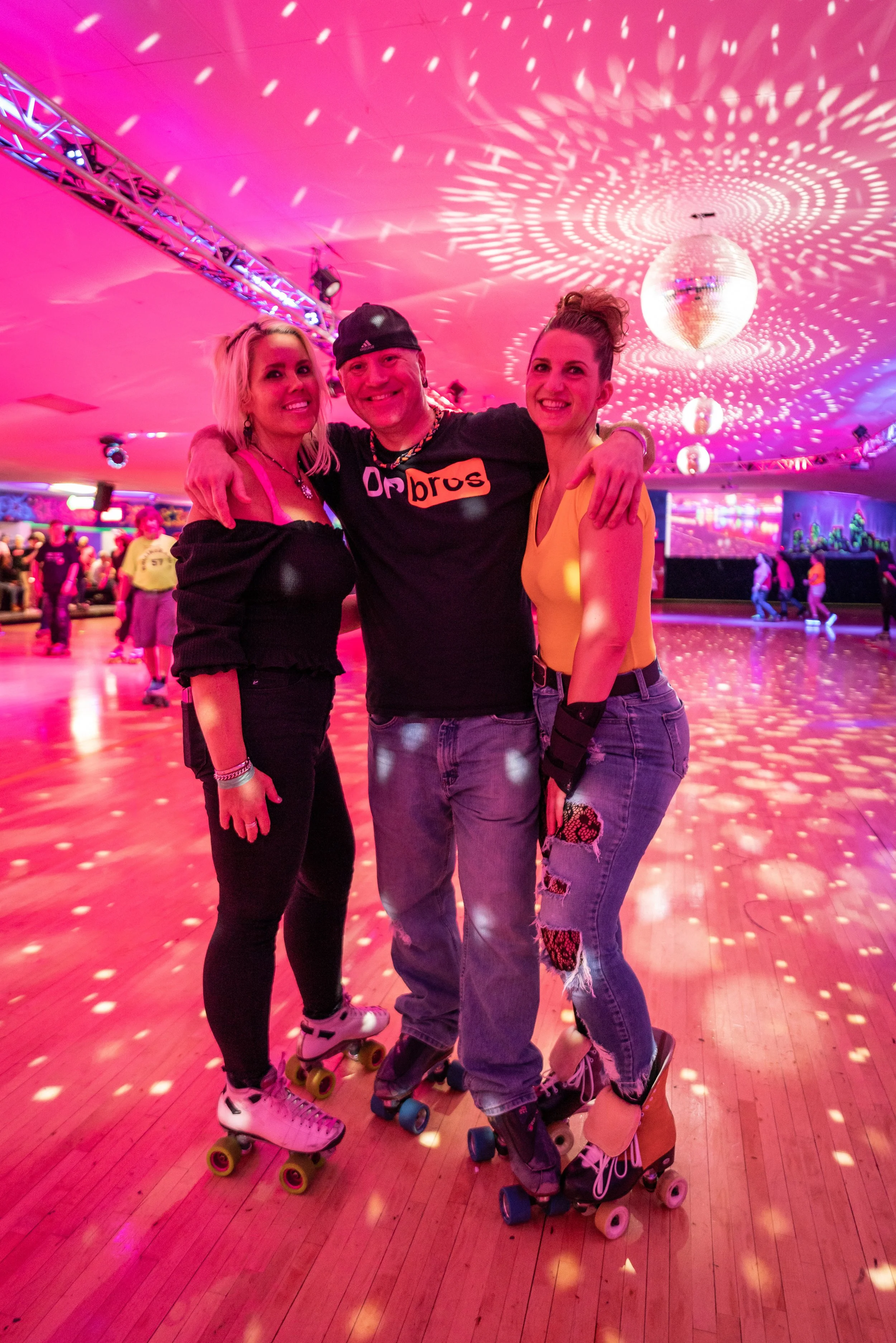 Three people smiling and posing together on roller skates in a roller skating rink with colorful lights and a disco ball overhead.