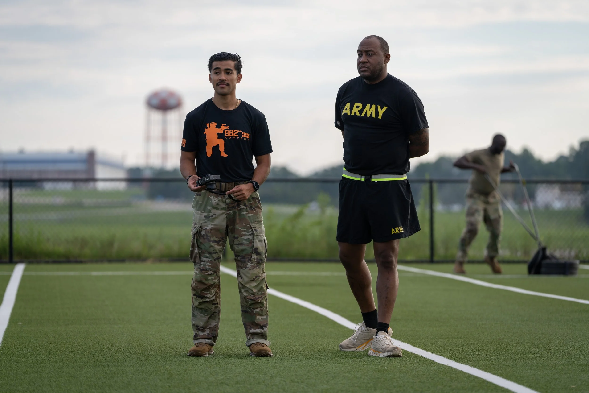 Two men standing on a sports field with a third person in the background. The man on the left wears camouflage pants, a black T-shirt with an orange graphic, and holds a remote control. The man on the right wears a black T-shirt with 'ARMY' printed o