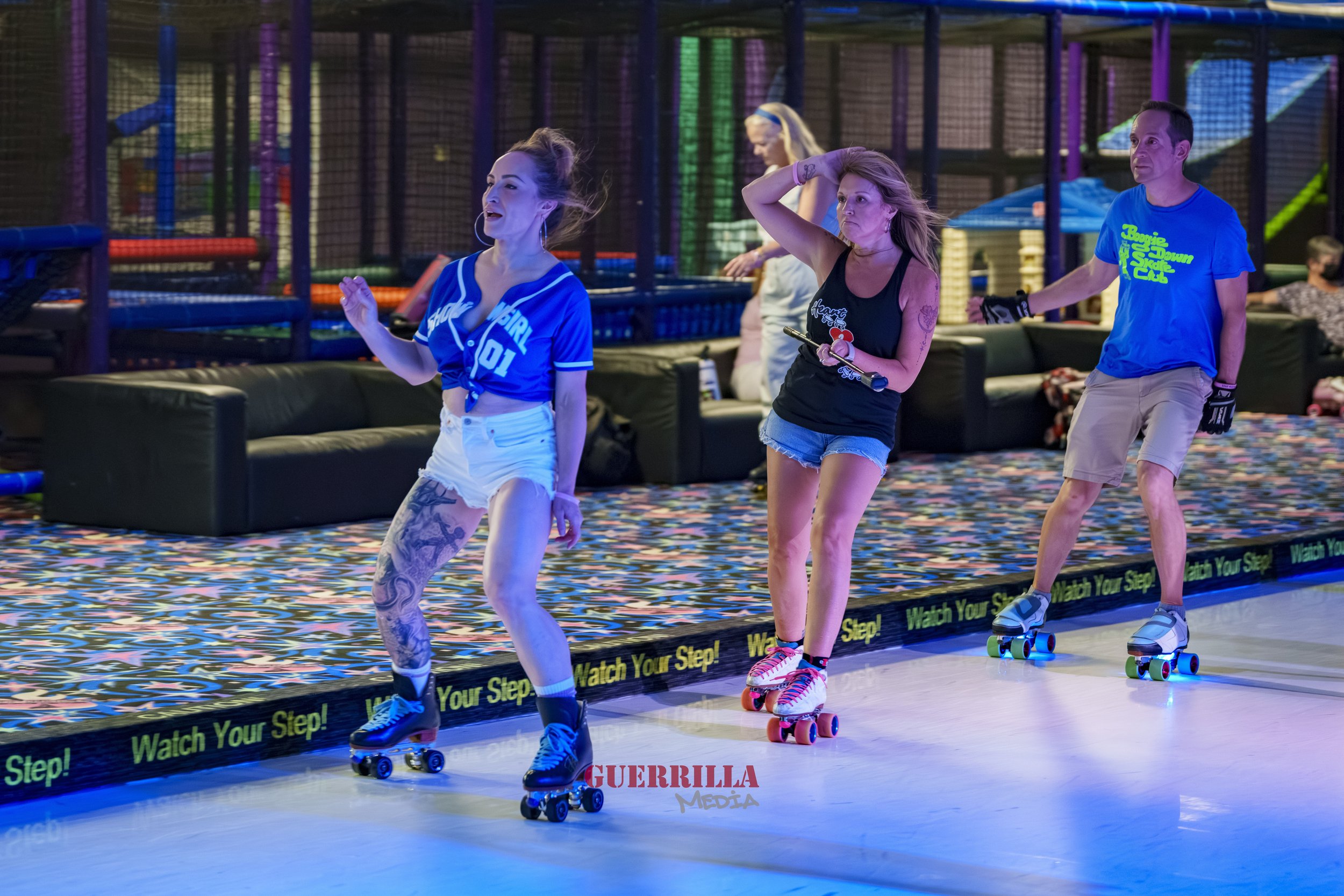 Three people roller skating at an indoor skating rink with colorful overhead lighting and padded barriers. One woman is wearing a blue baseball jersey and white shorts, another woman is wearing a black tank top and denim shorts, and a man is in a bri