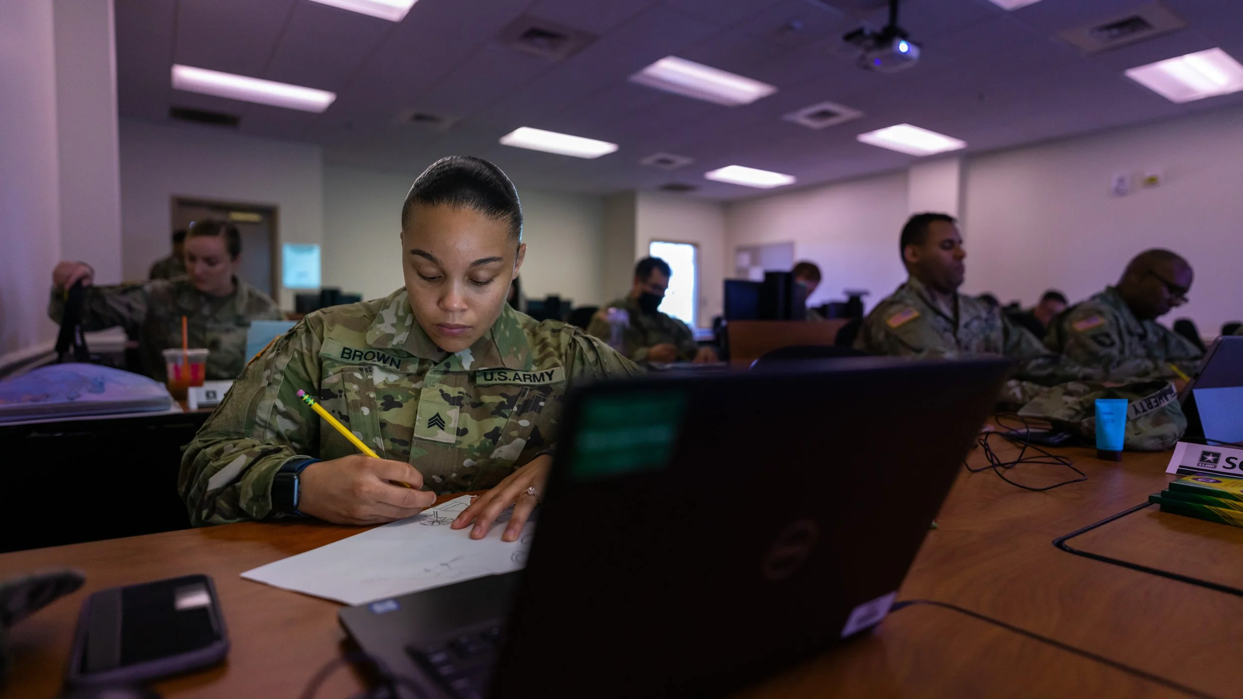 Military personnel in uniform seated at a conference table using laptops and taking notes in a classroom setting.