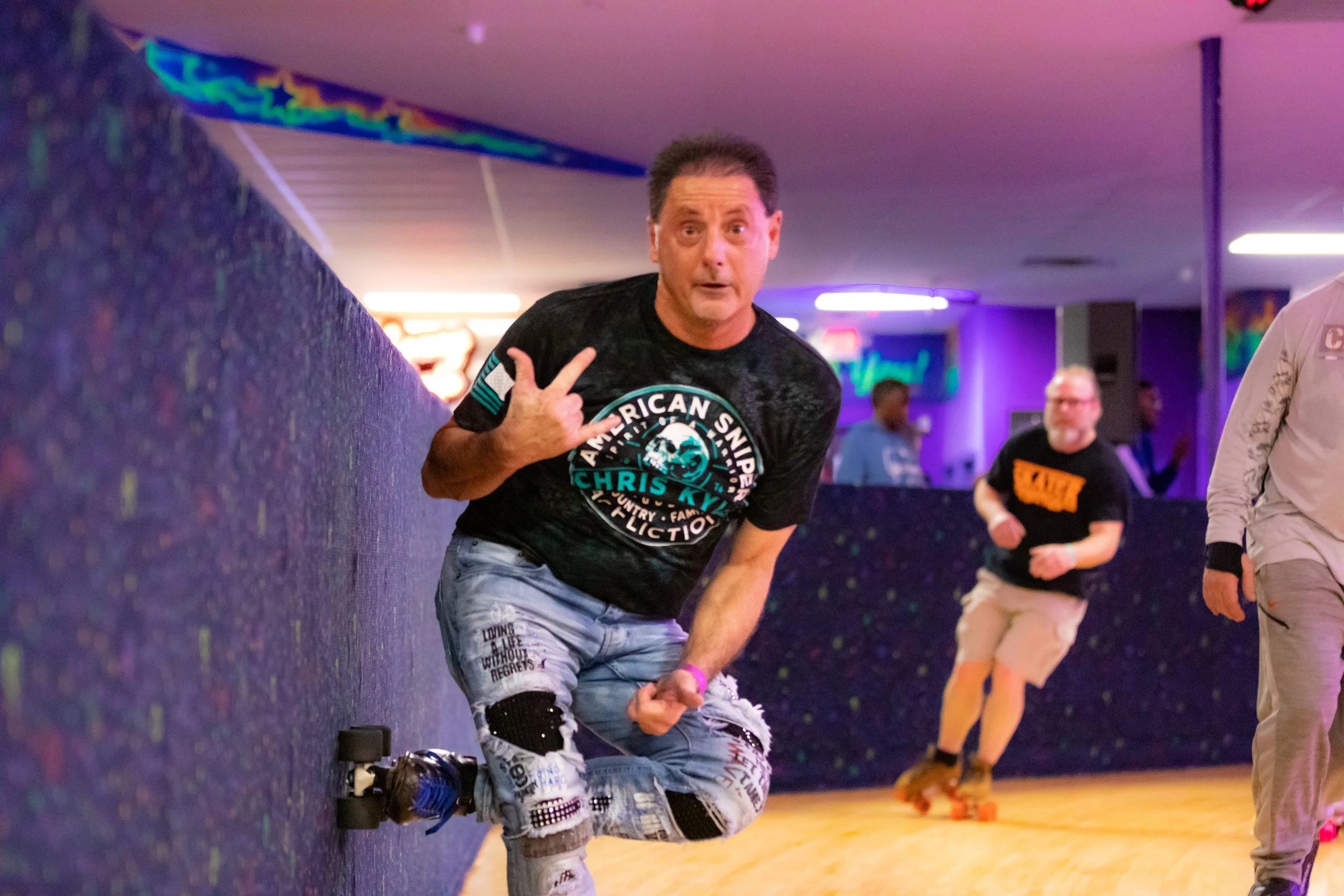 A man roller skating indoors with a surprised expression, wearing a black T-shirt and ripped jeans, making a hand gesture with his left hand, with other skaters and colorful neon lights in the background.
