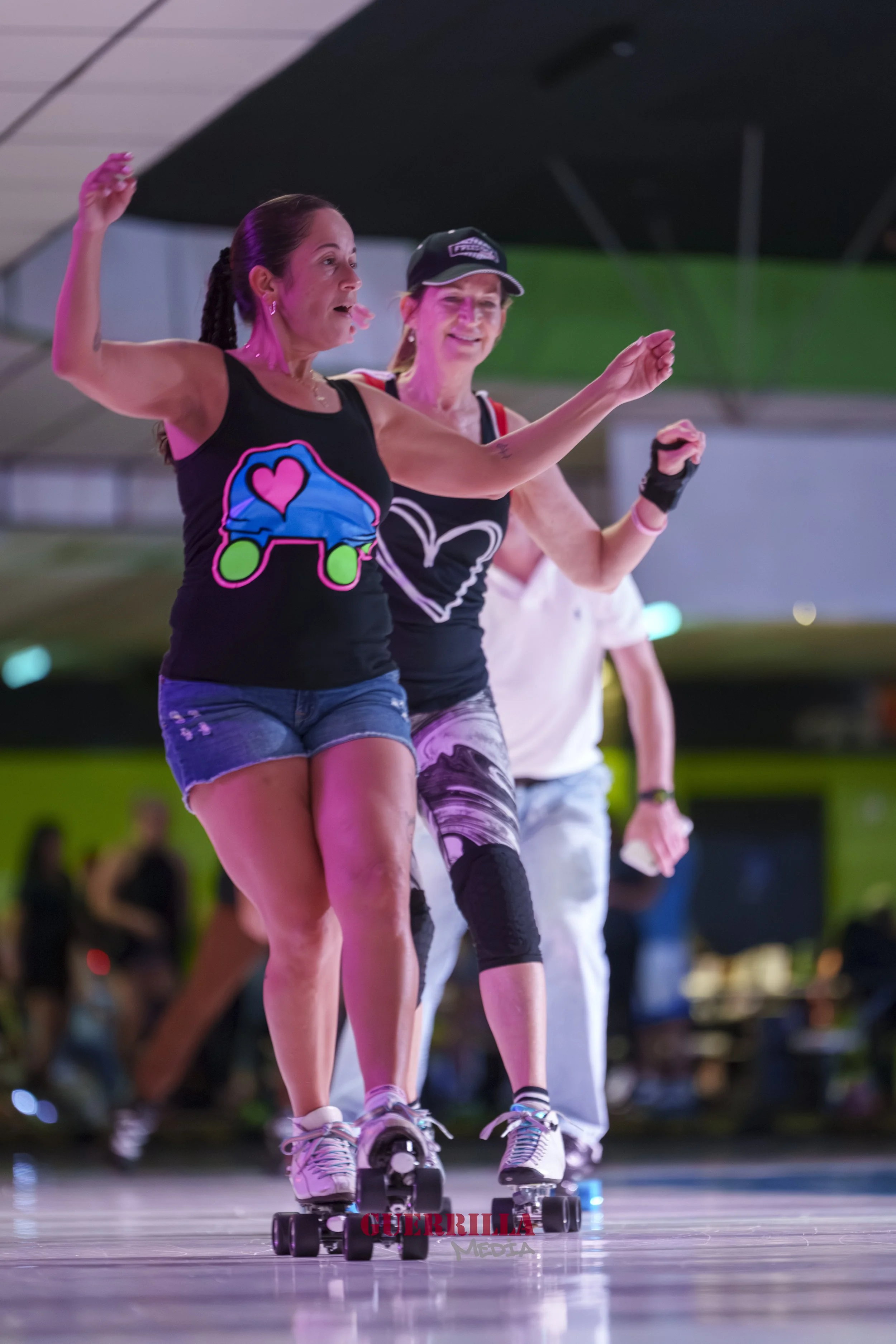 Two women roller skating indoors, one leading and the other following, both smiling and wearing athletic clothing with logos.