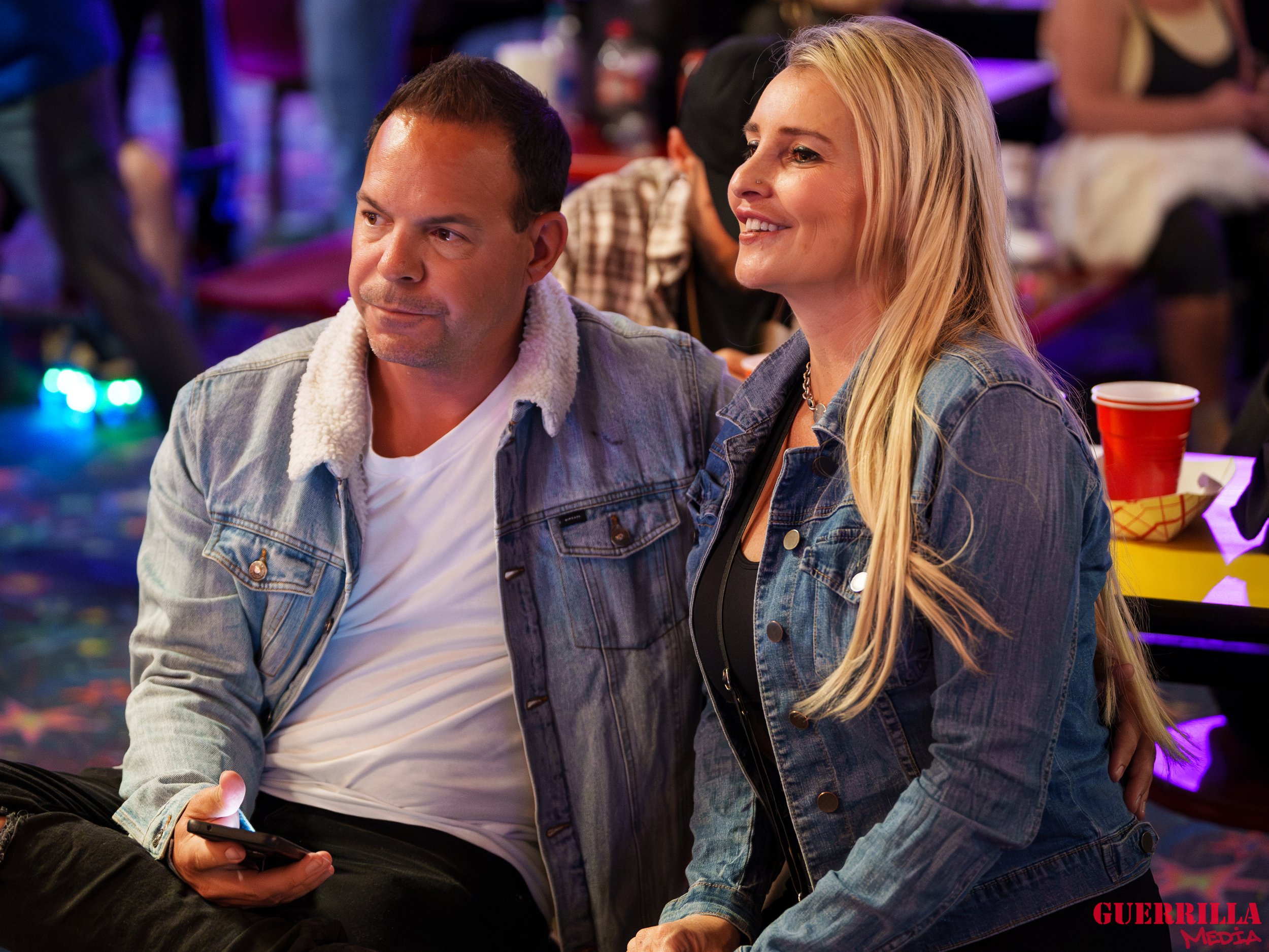 A man and woman sitting at an arcade, the man holding a smartphone, both wearing denim jackets, with colorful arcade lights in the background.