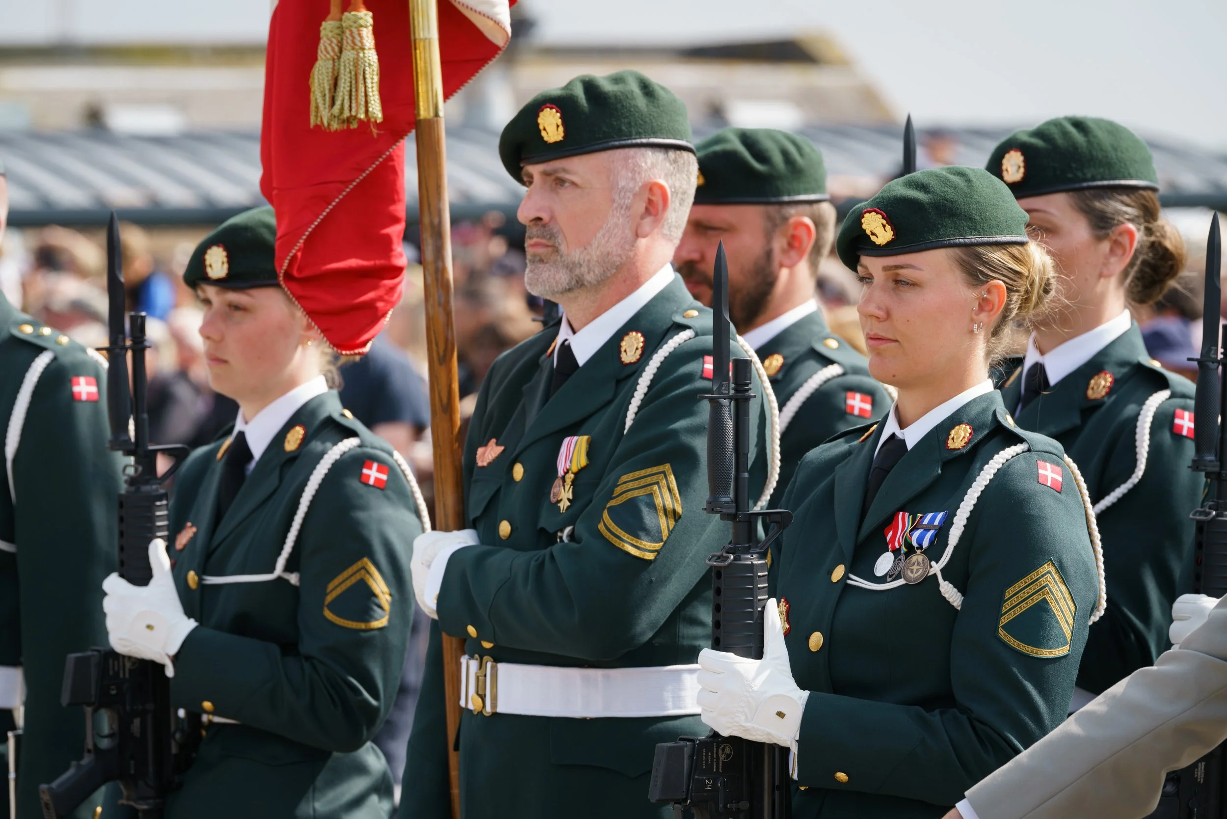 Danish military personnel in uniform standing in formation, participating in a ceremonial event, with some holding rifles and wearing medals, while others hold a flag.