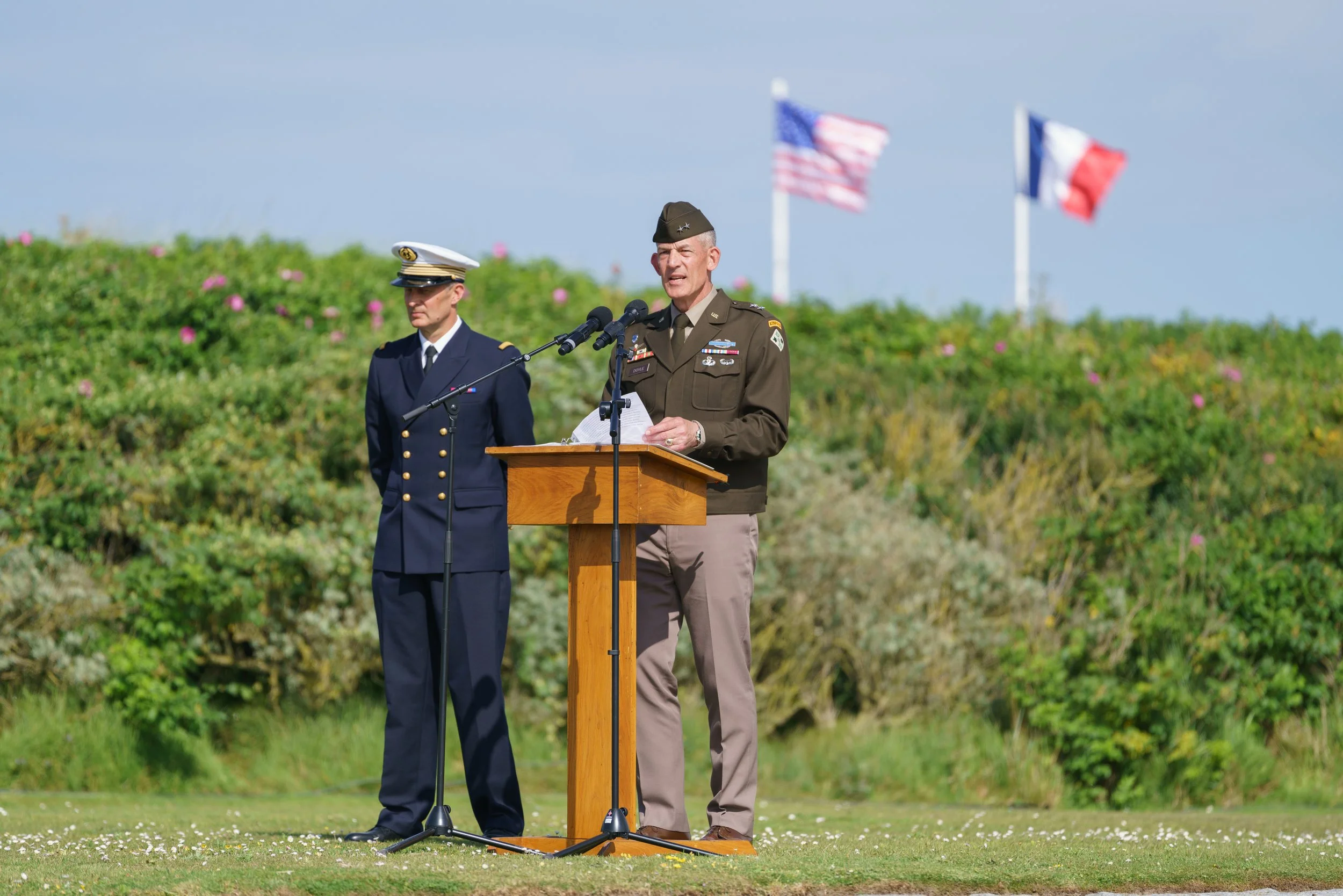 Military officer speaking at a podium during an outdoor ceremony, with a woman in a navy uniform standing beside him, and American and French flags in the background.
