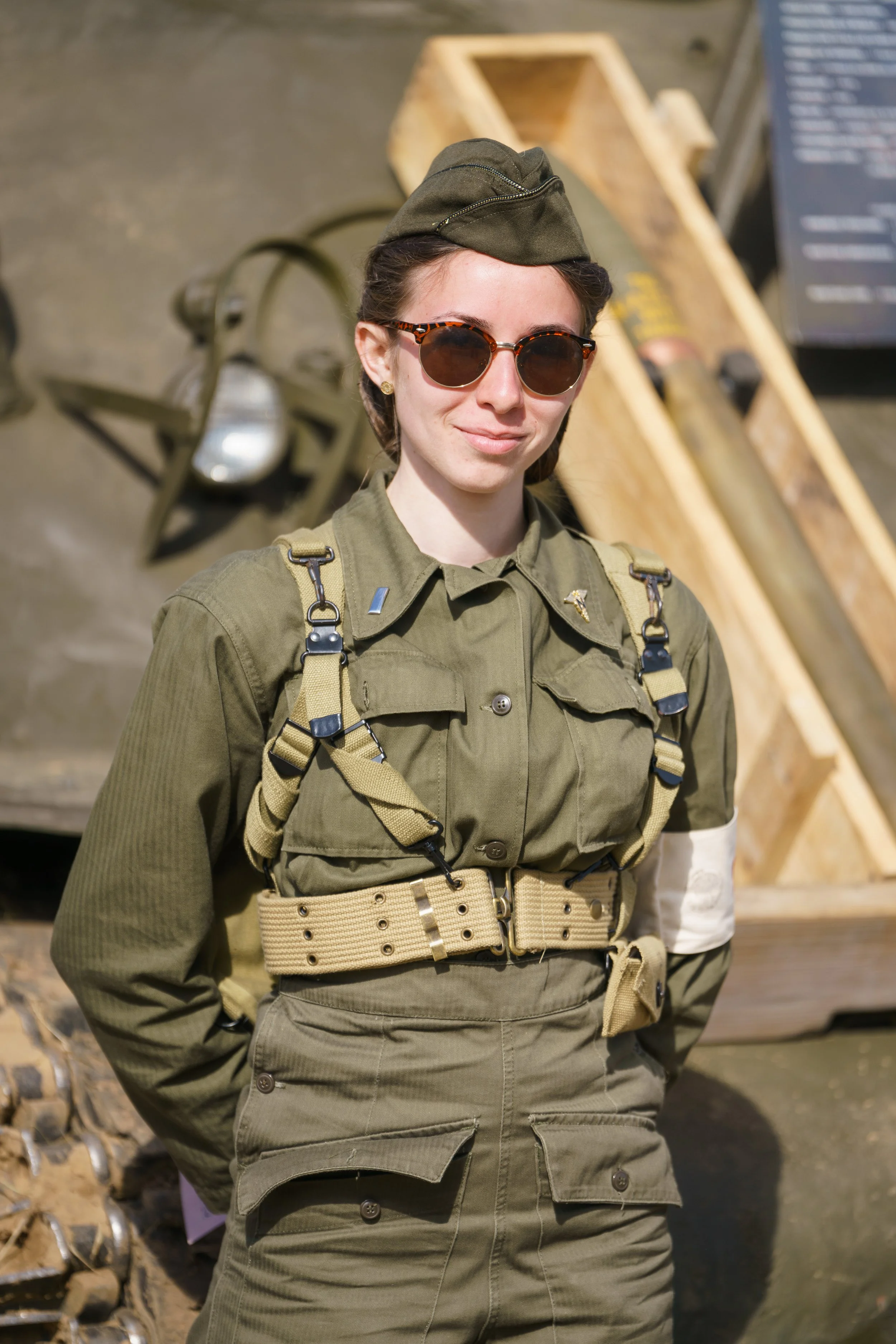 A woman dressed in military uniform with tactical gear, wearing sunglasses and a cap, standing outdoors with tools and wooden crates in the background.