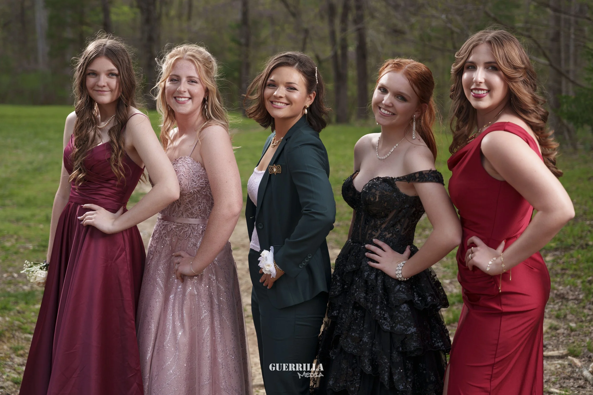 Five young women dressed in formal gowns and a blazer, standing side by side outdoors in a forested area.