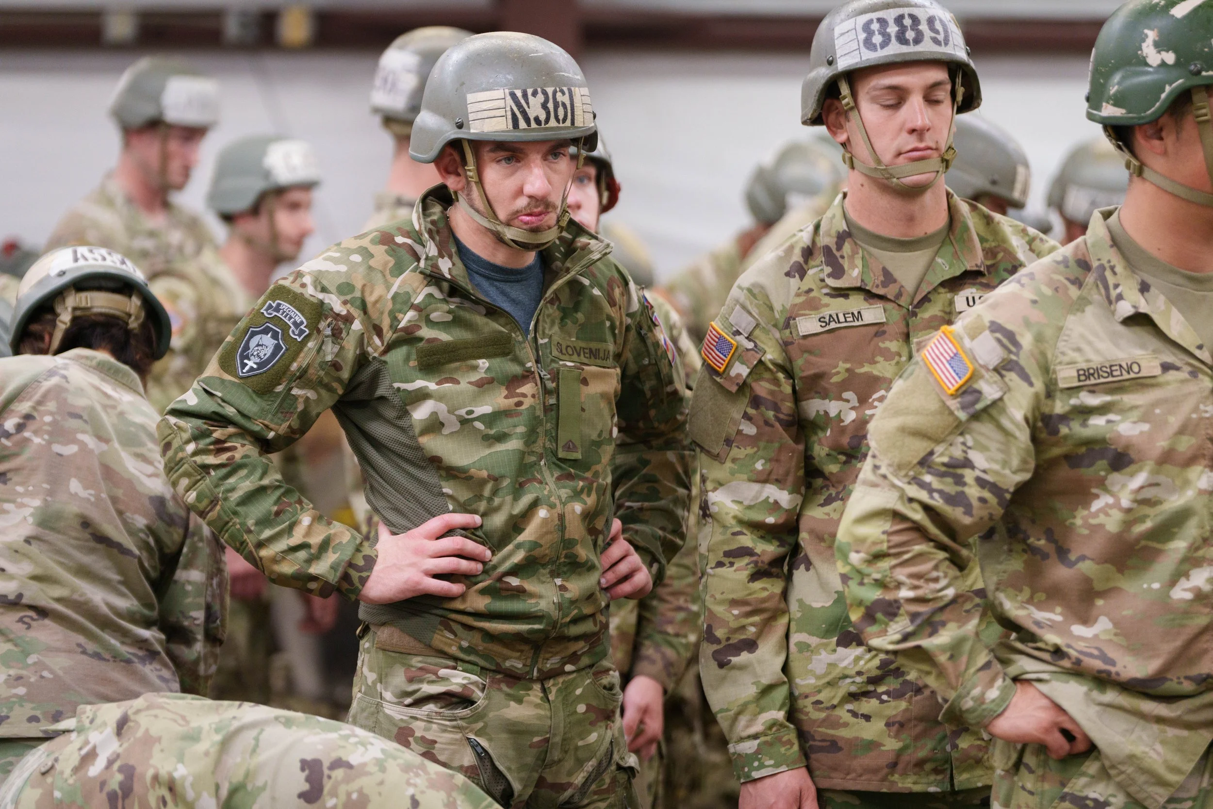 Group of soldiers in camouflage uniforms and helmets standing in formation, with some looking serious and others with eyes closed, inside a military facility.