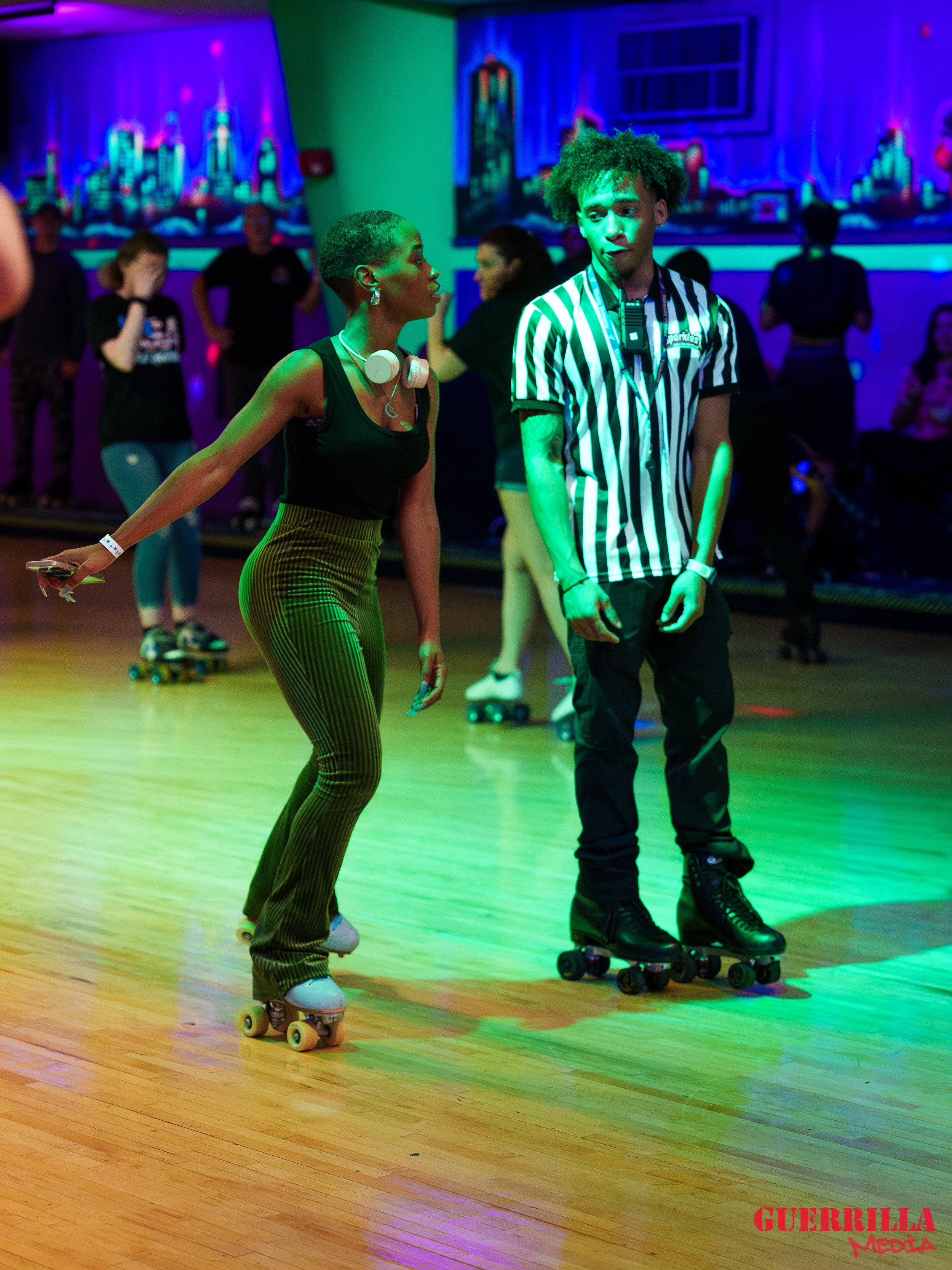 Two young adults roller skating at an indoor roller rink with colorful neon lights, one woman and one man, engaging in conversation.