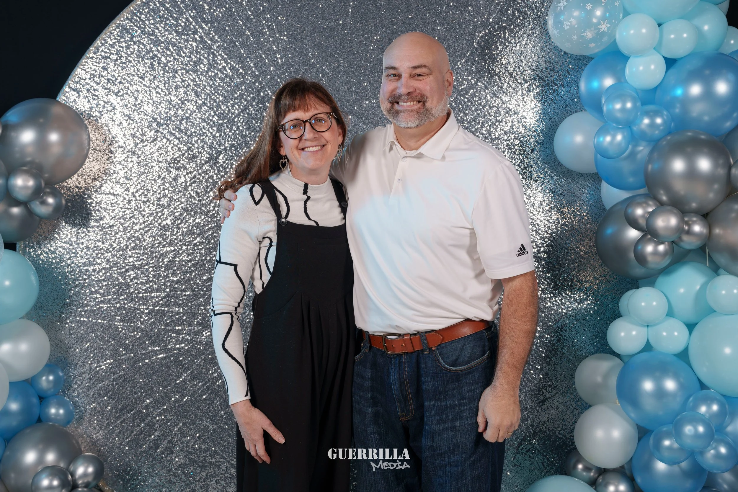 A smiling couple posing in front of a shiny silver backdrop with blue and silver balloons on either side.