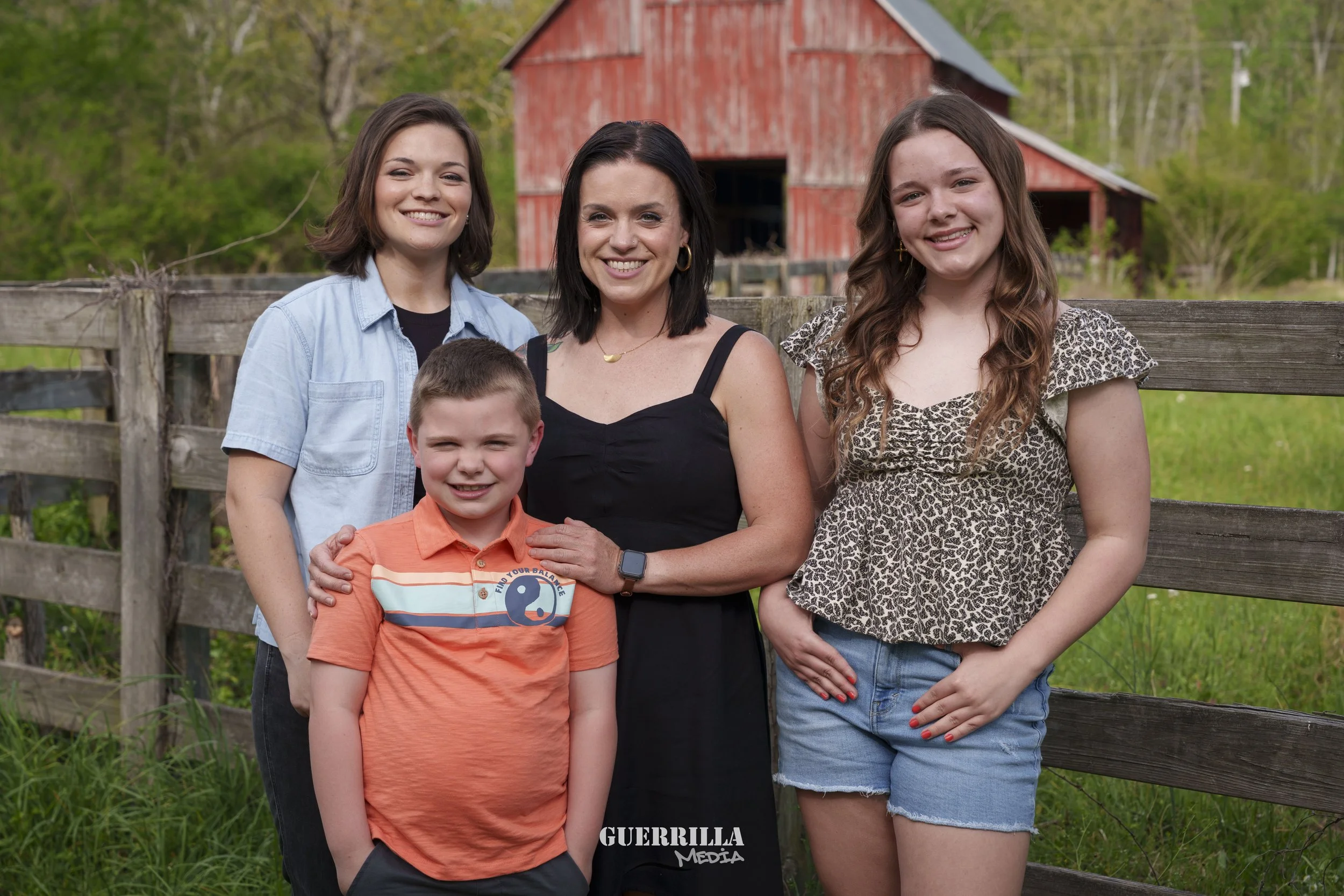A woman with black hair wearing a black dress stands outside with three children near a wooden fence with a red barn and green trees in the background.