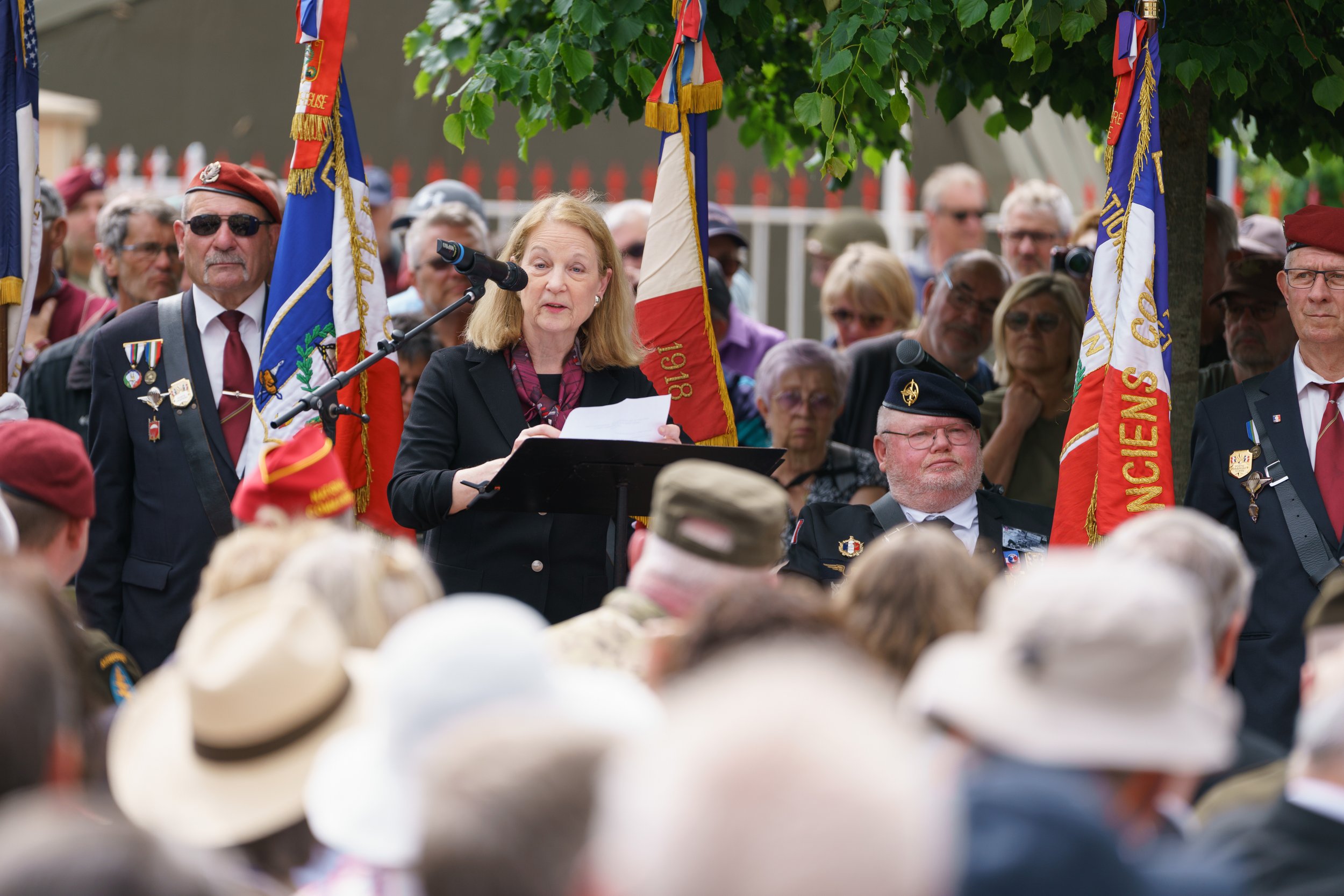 A woman is speaking at a public event with a microphone, surrounded by people including men in military or veteran uniforms and flags, under a tree.