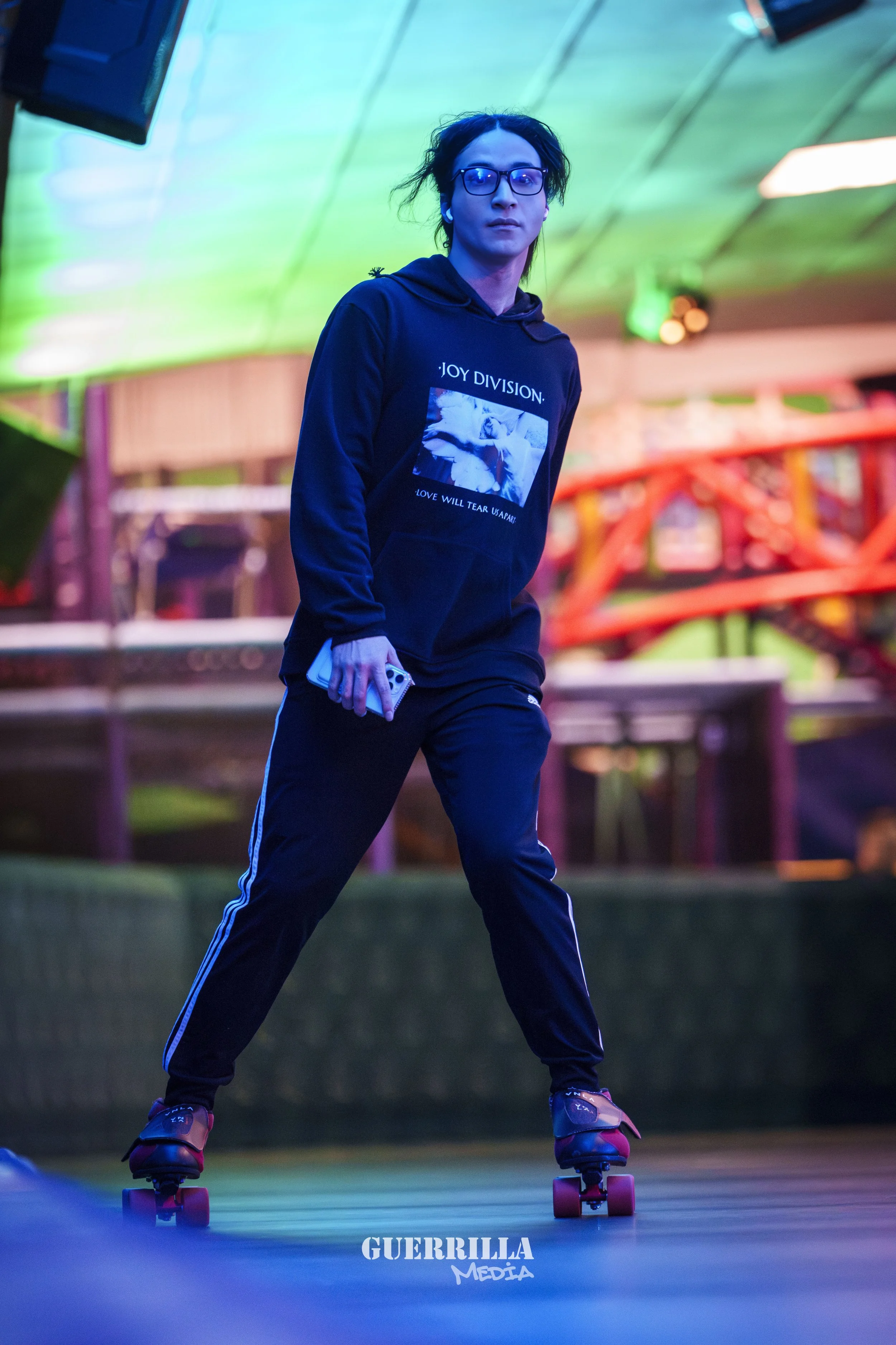 A person riding a skateboard indoors under colorful lighting, wearing glasses, a black Joy Division hoodie, and black pants.