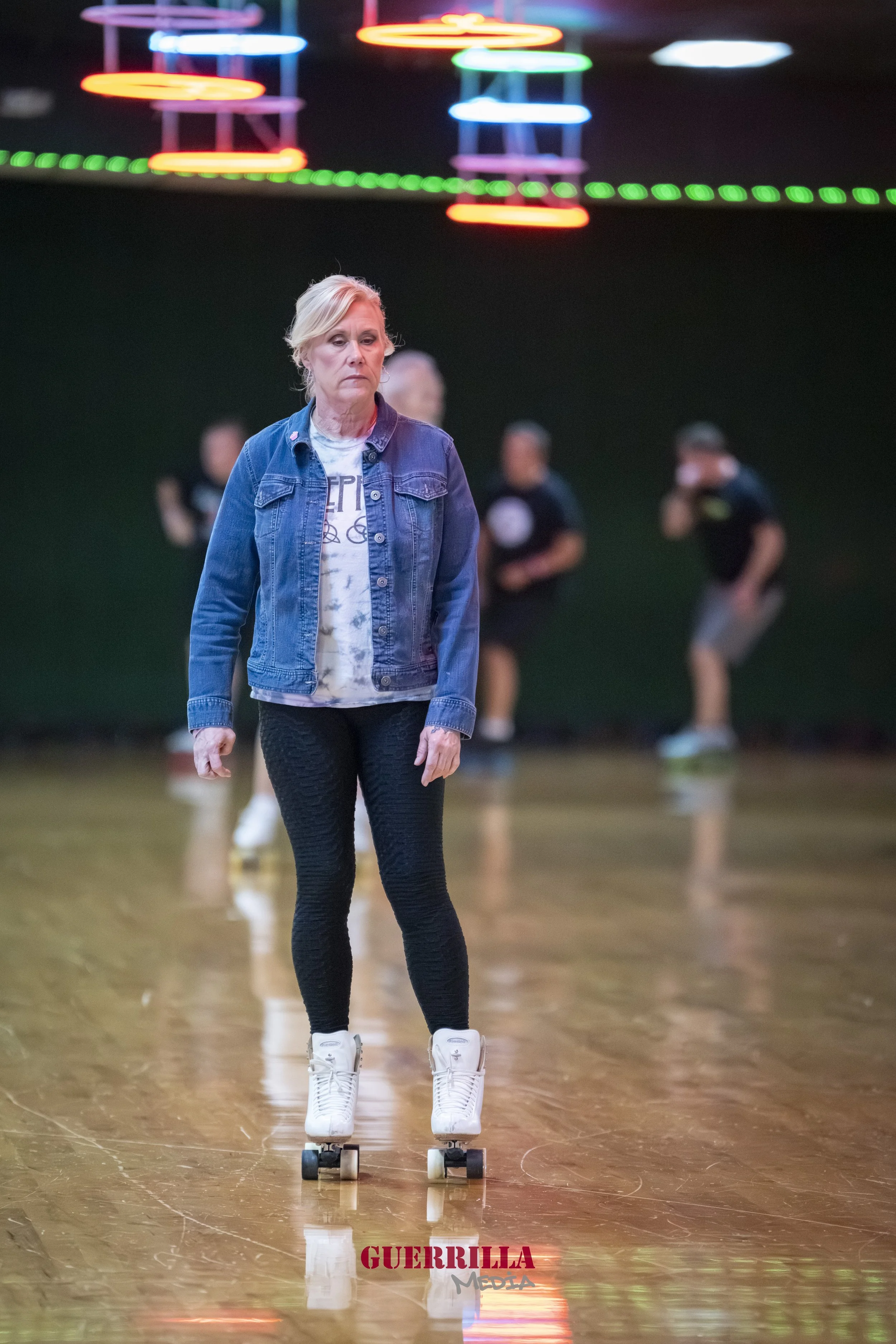A woman standing on roller skates inside a roller skating rink, with three young men skating in the background, and colorful neon lights overhead.
