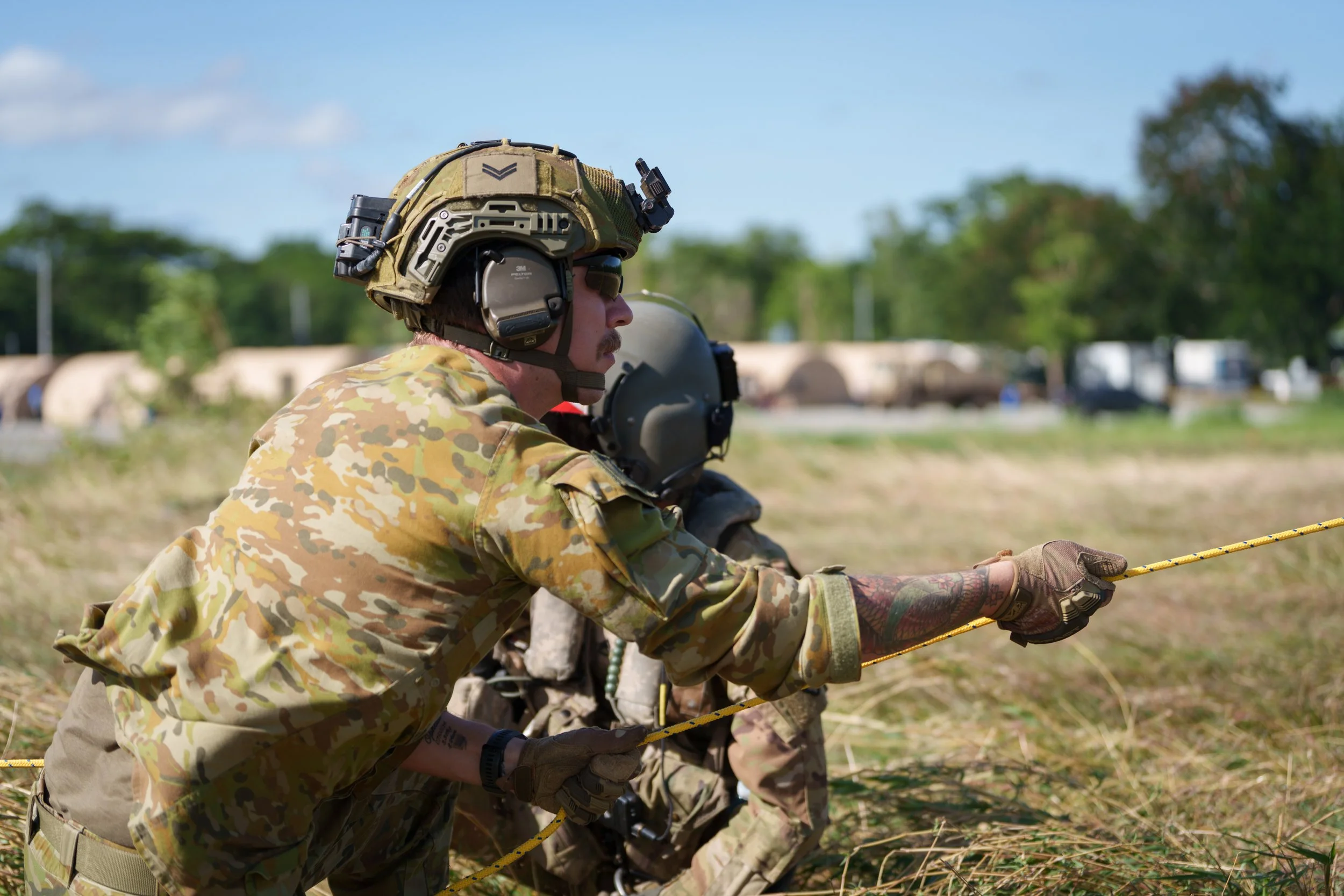A soldier in camouflage uniform and tactical gear is holding a yellow rope during a training exercise in an open field.