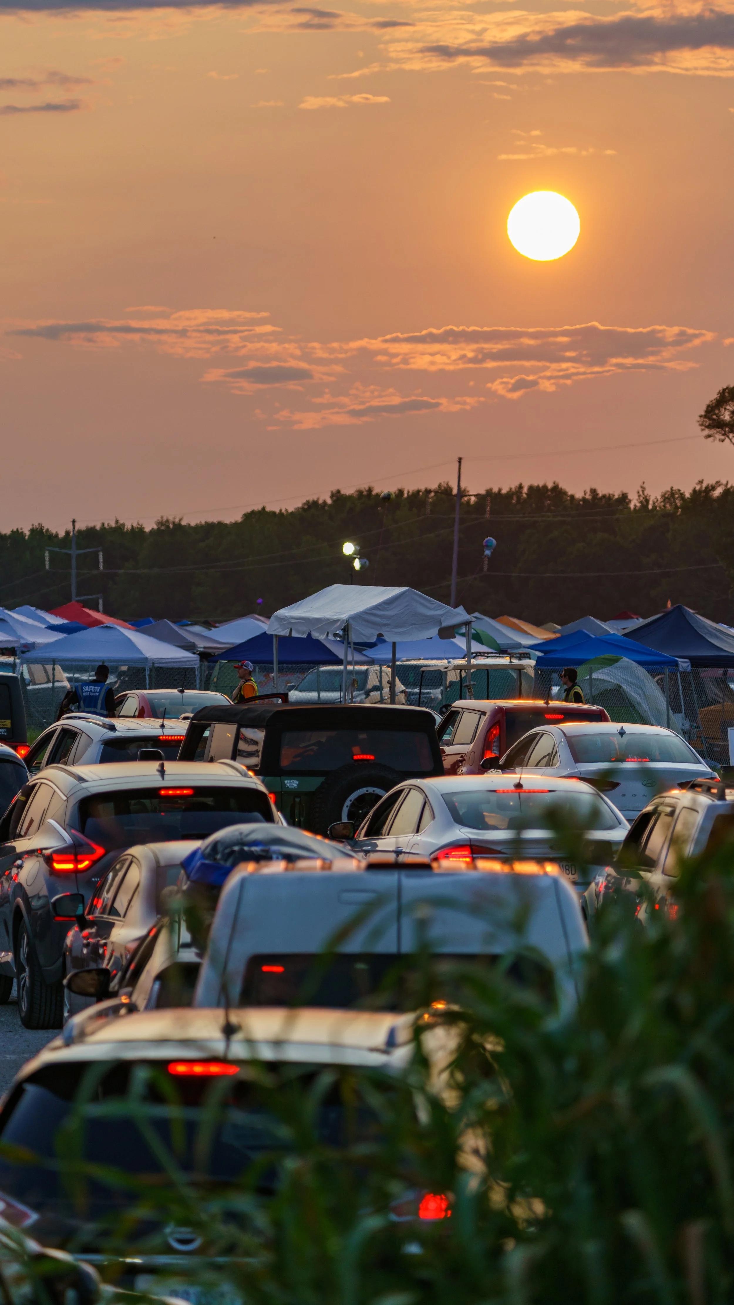 Sunset over a parking lot with numerous parked cars and tents, with people walking and trees in the background.