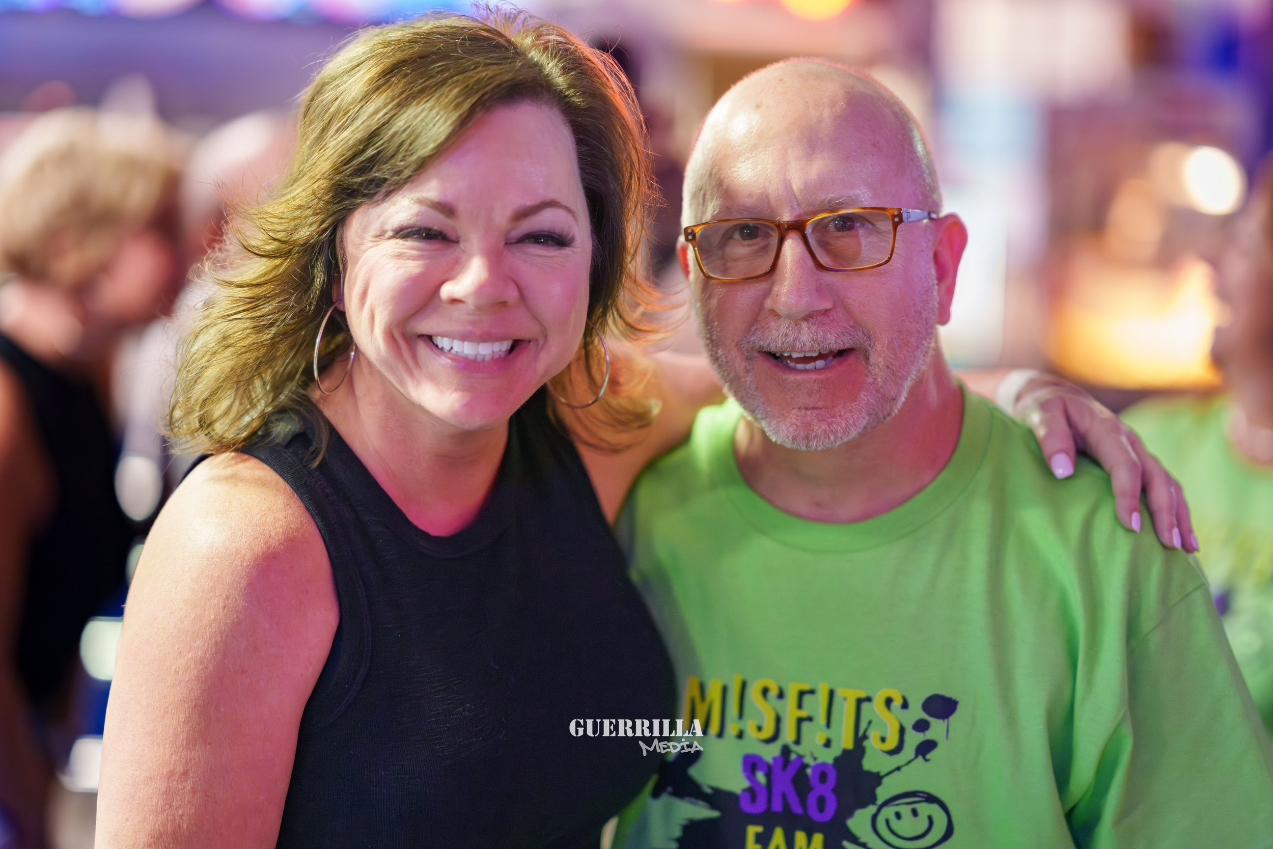A smiling woman with shoulder-length wavy hair and hoop earrings stands next to a man with a beard and glasses, both in a colorful social setting.