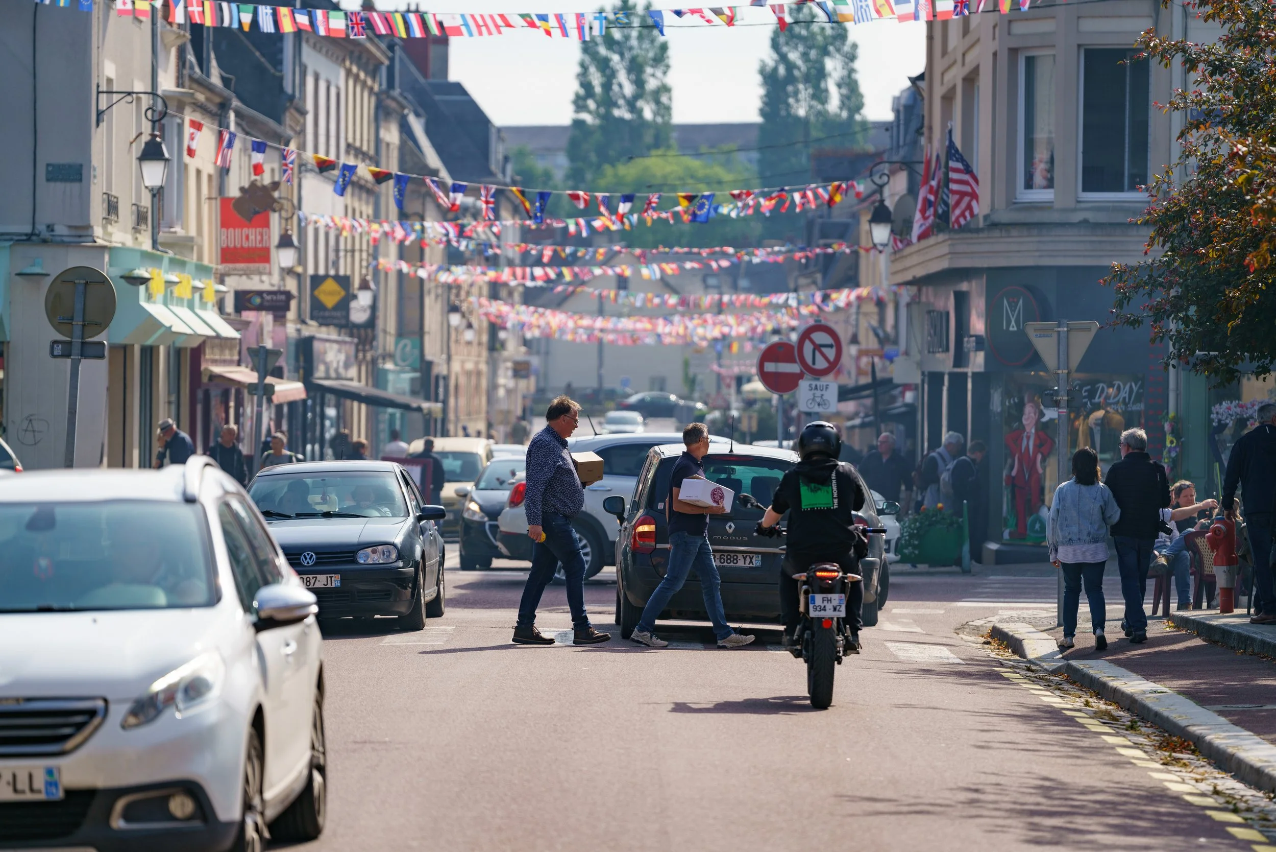 A lively street scene decorated with hanging flag banners, featuring pedestrians crossing and walking along the sidewalks, cars parked and driving, and a motorcycle rider in front, during daytime.