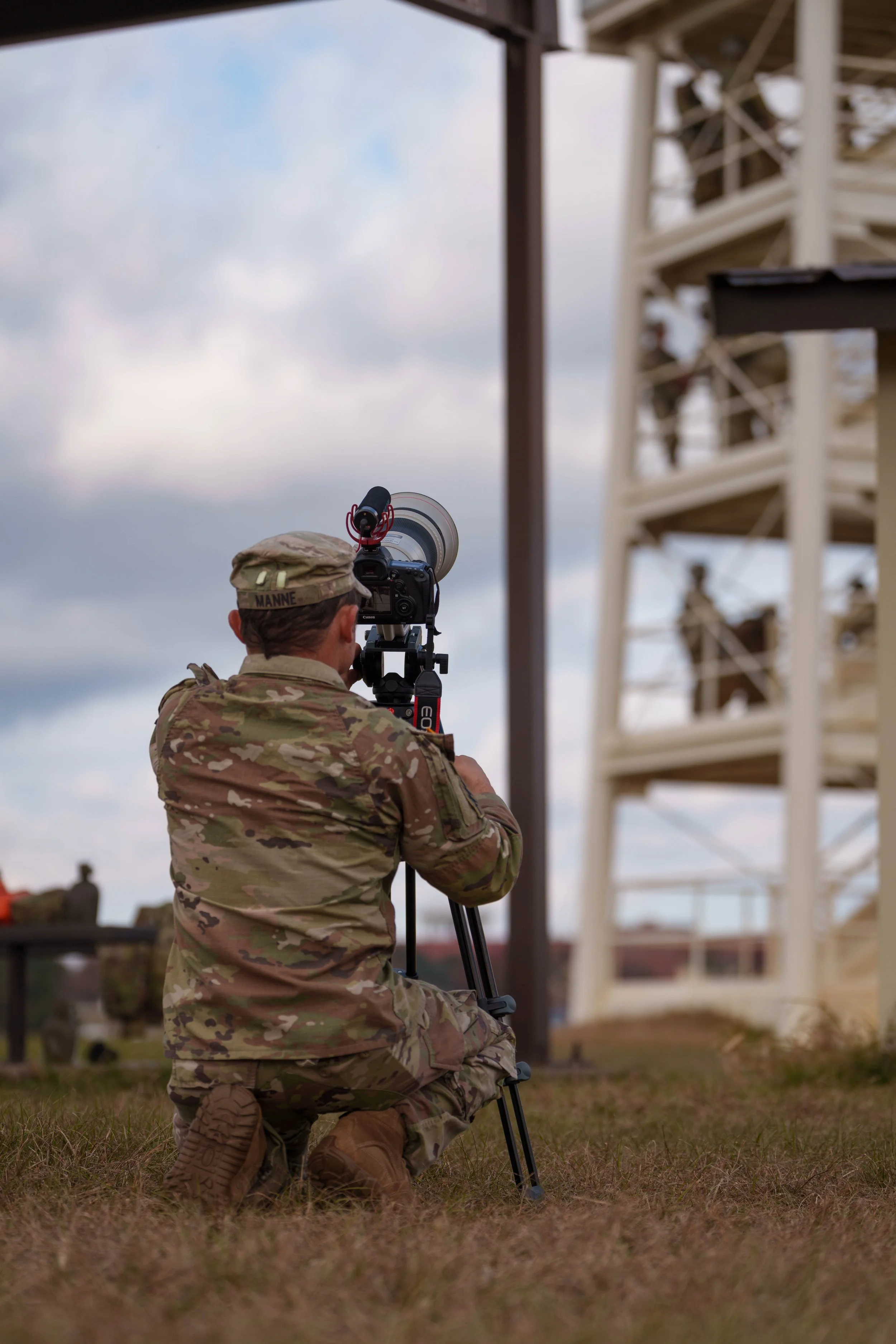 A soldier in camouflage uniform kneeling on grass and operating a camera on a tripod, with a structural tower and clouds in the background.