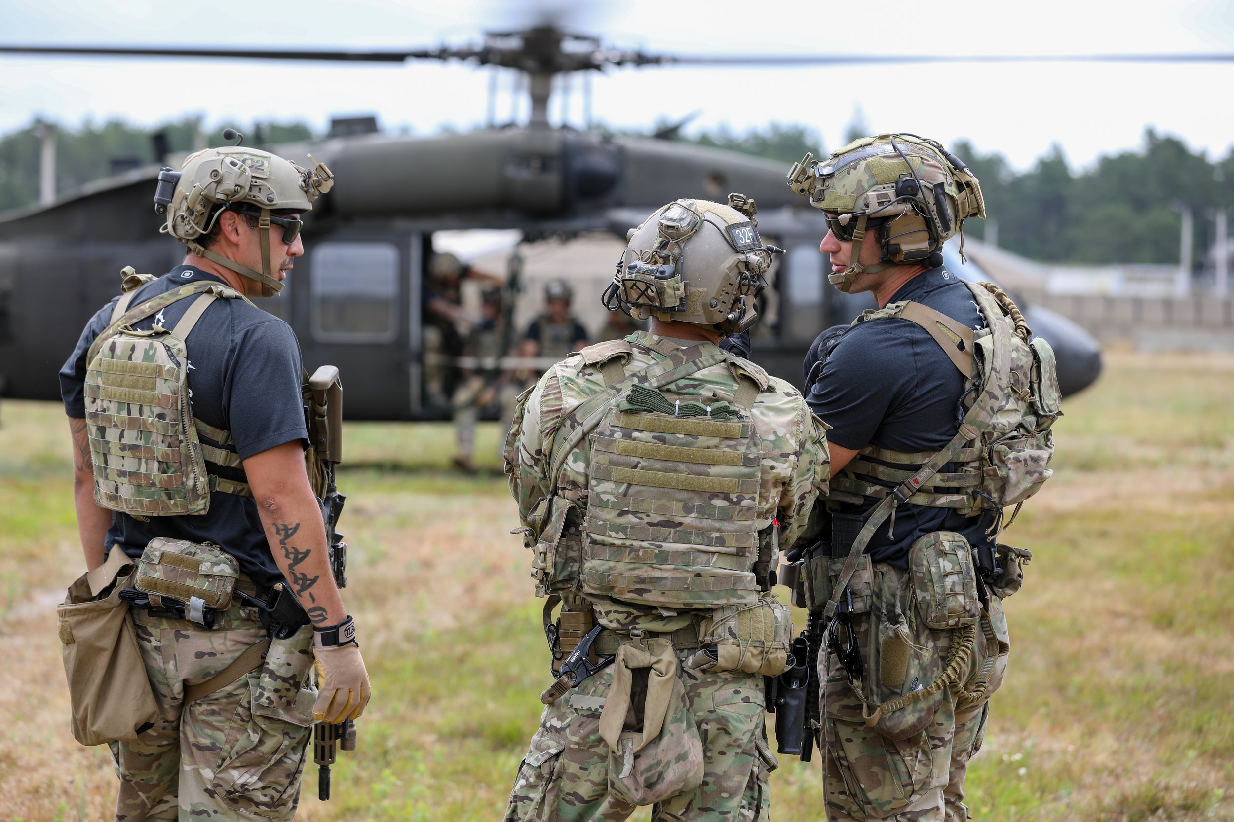 Three soldiers in camouflage military gear standing in front of a black helicopter, having a discussion outdoors.