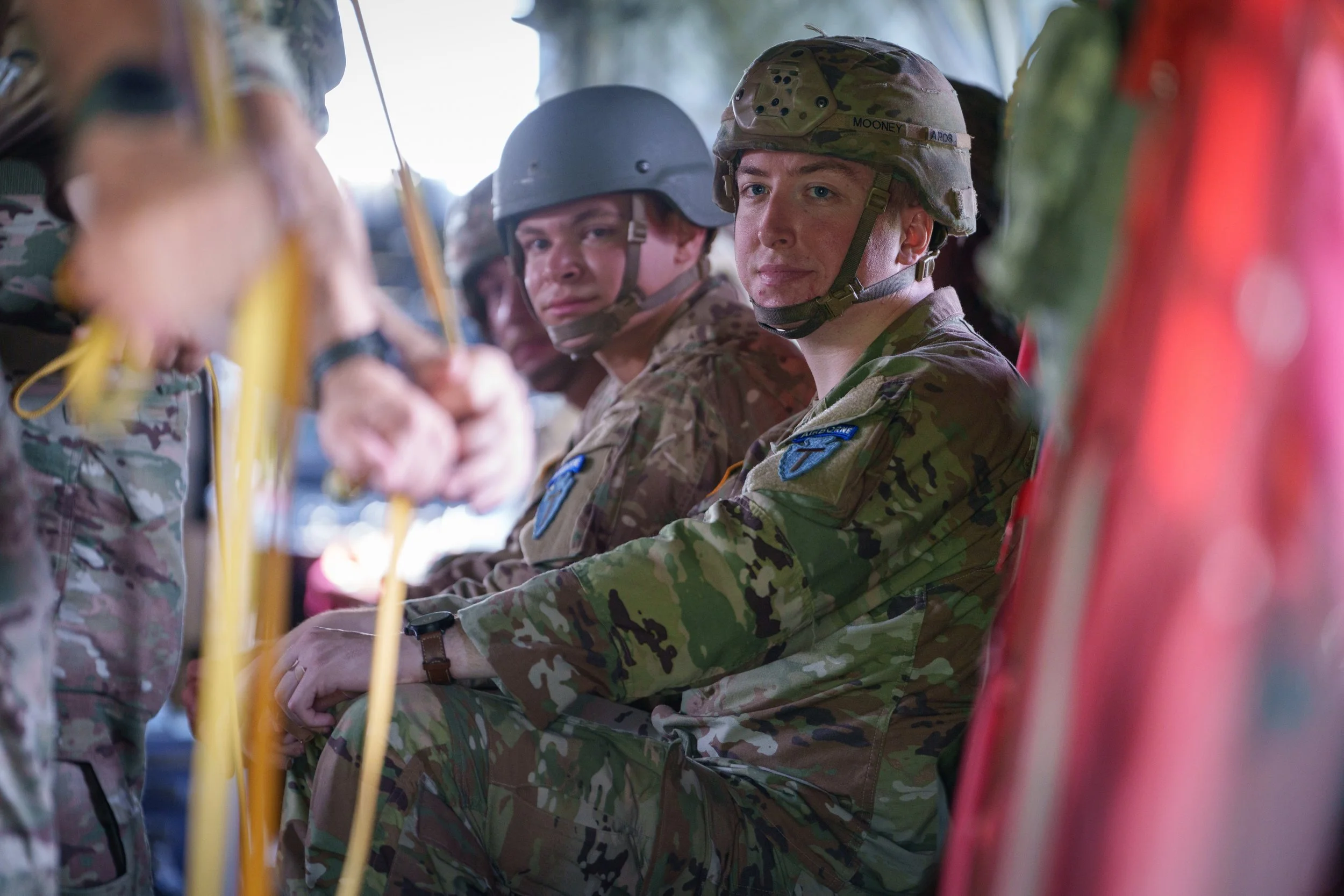 Three soldiers in camouflage uniforms sitting in a row on a military aircraft, with serious expressions, wearing helmets, and surrounded by other soldiers and equipment.
