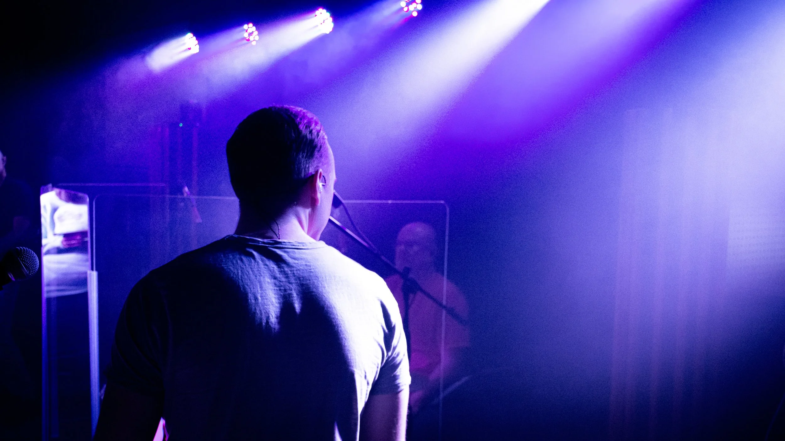 Back of a person performing on stage with purple and white lighting, microphones, and other performers in the background.